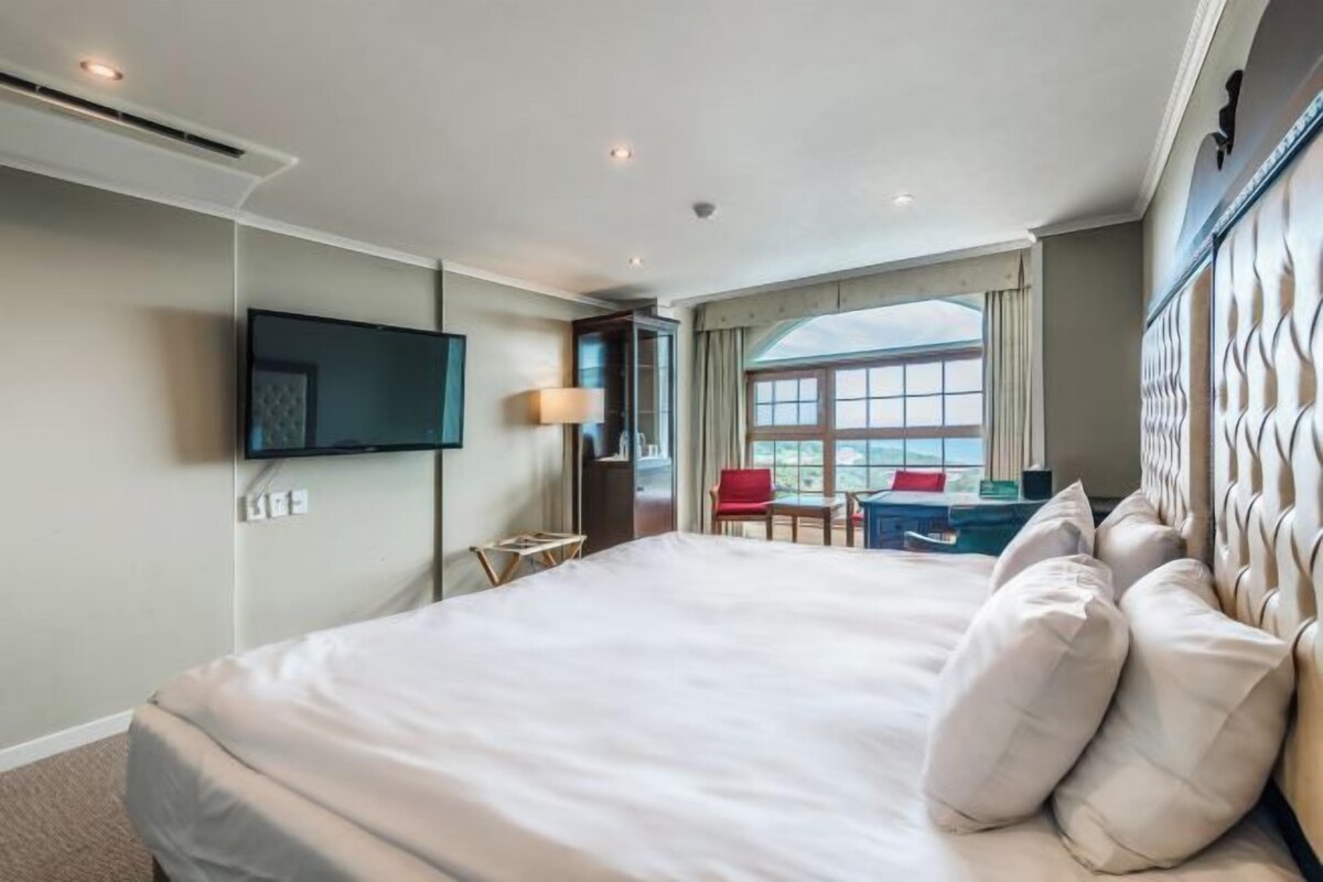 A hotel room with a large bed, a TV mounted on the wall, a desk with a chair, and a window with a view.