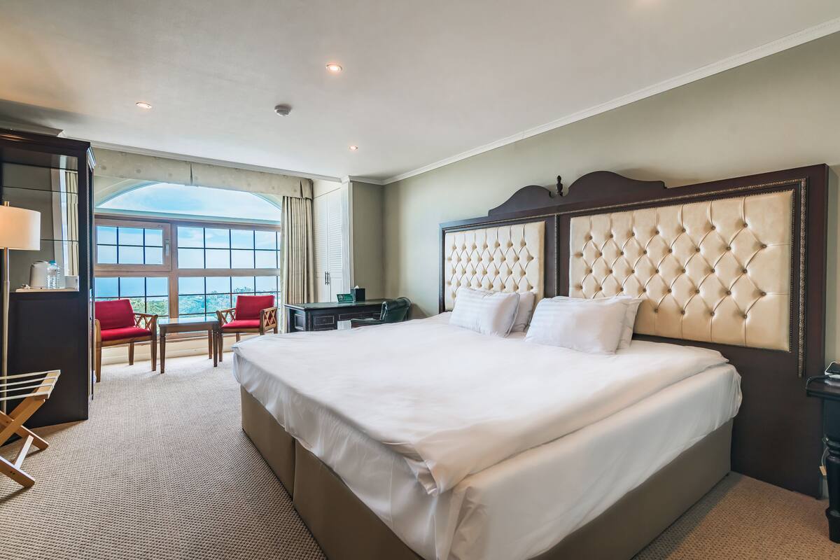 A spacious hotel room with a large bed, a desk, and a chair. There is a window with a view of the sky.