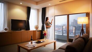 Habitación de hotel con televisión, una mesa de comedor con una botella y un plato de fruta, un sofá, un ventanal con vistas a la ciudad y una lámpara.
