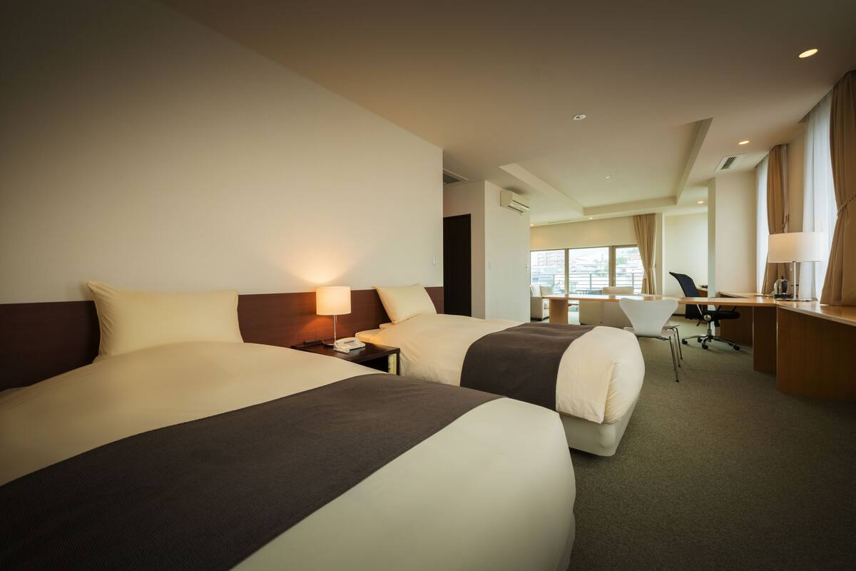 A hotel room with two beds, a desk, a chair, and a large window.