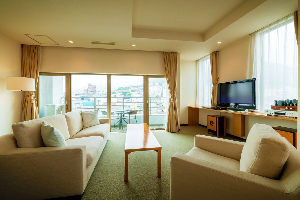 A modern hotel room with a sofa, a coffee table, a TV, and a balcony with a city view.
