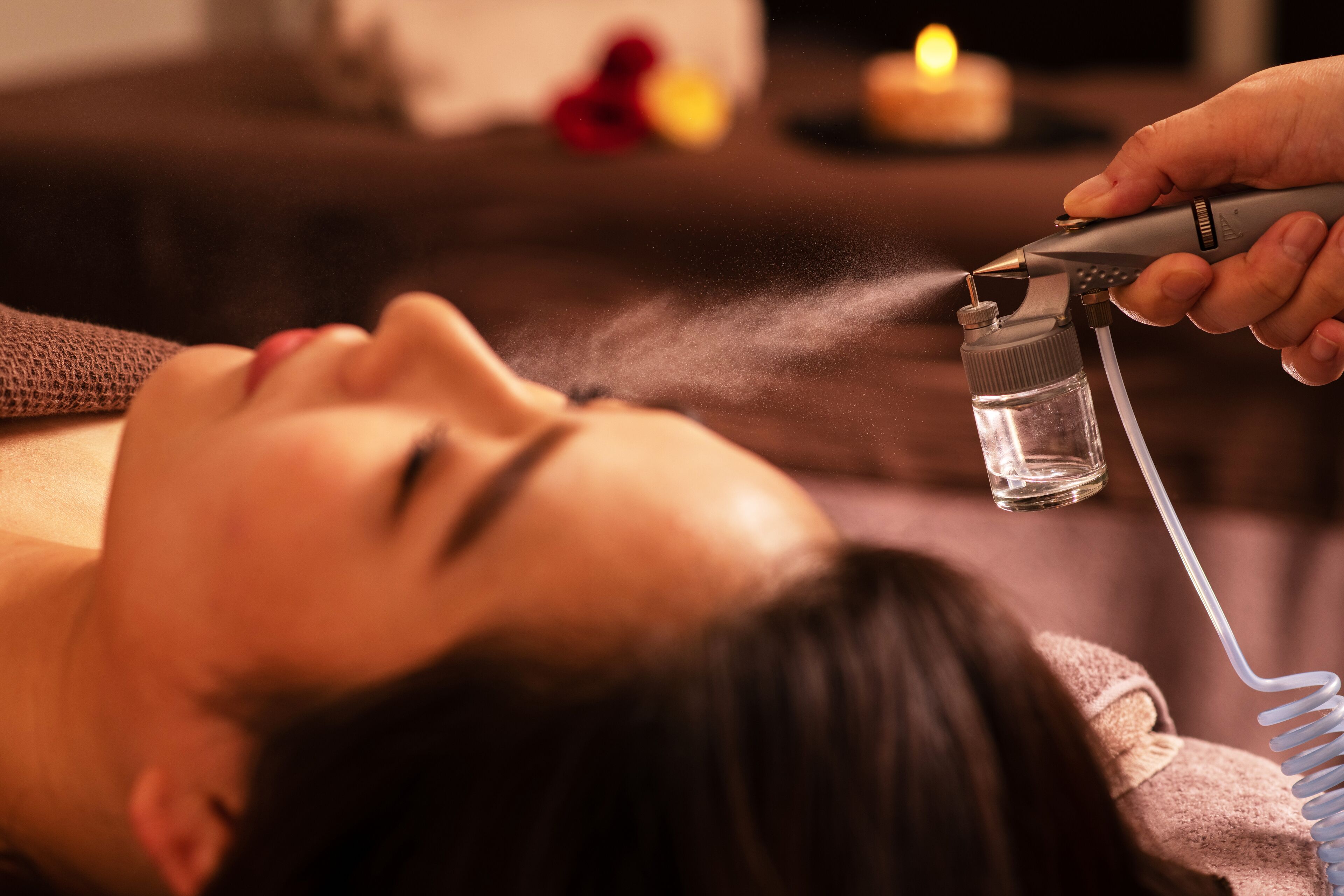 Eine Person liegt entspannt, während ein Gerät Nebel abgibt, möglicherweise für eine Spa-Behandlung, in einem Raum mit Kerze und unscharfem Hintergrund.