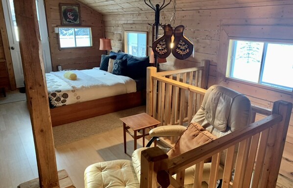 Una habitación tipo cabaña acogedora con una cama de madera, una mesita pequeña, un sillón con cojines y una lámpara colgante.