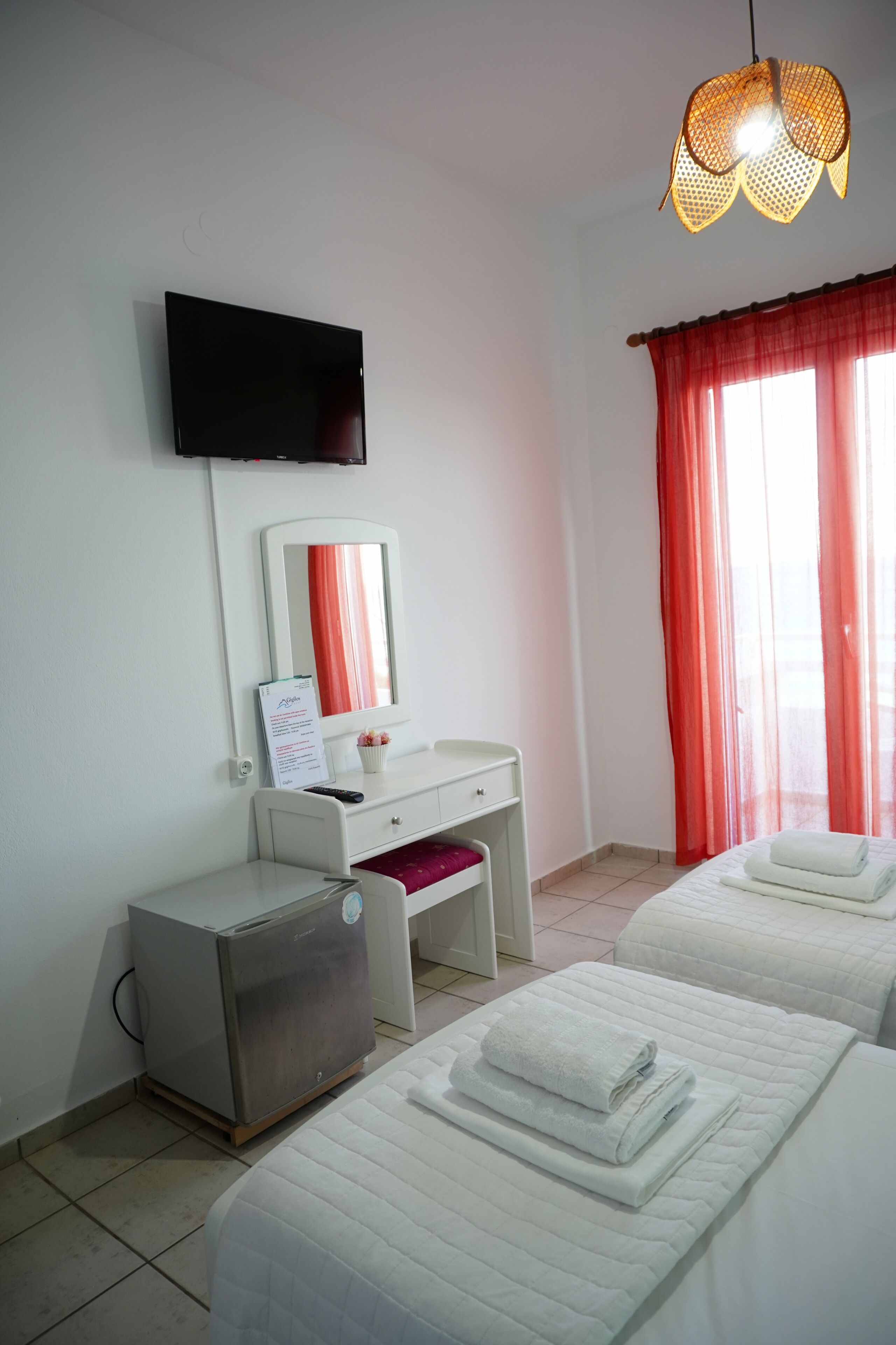 A hotel room with a flat-screen TV, a desk with a mirror, two beds with towels, and a ceiling light fixture.