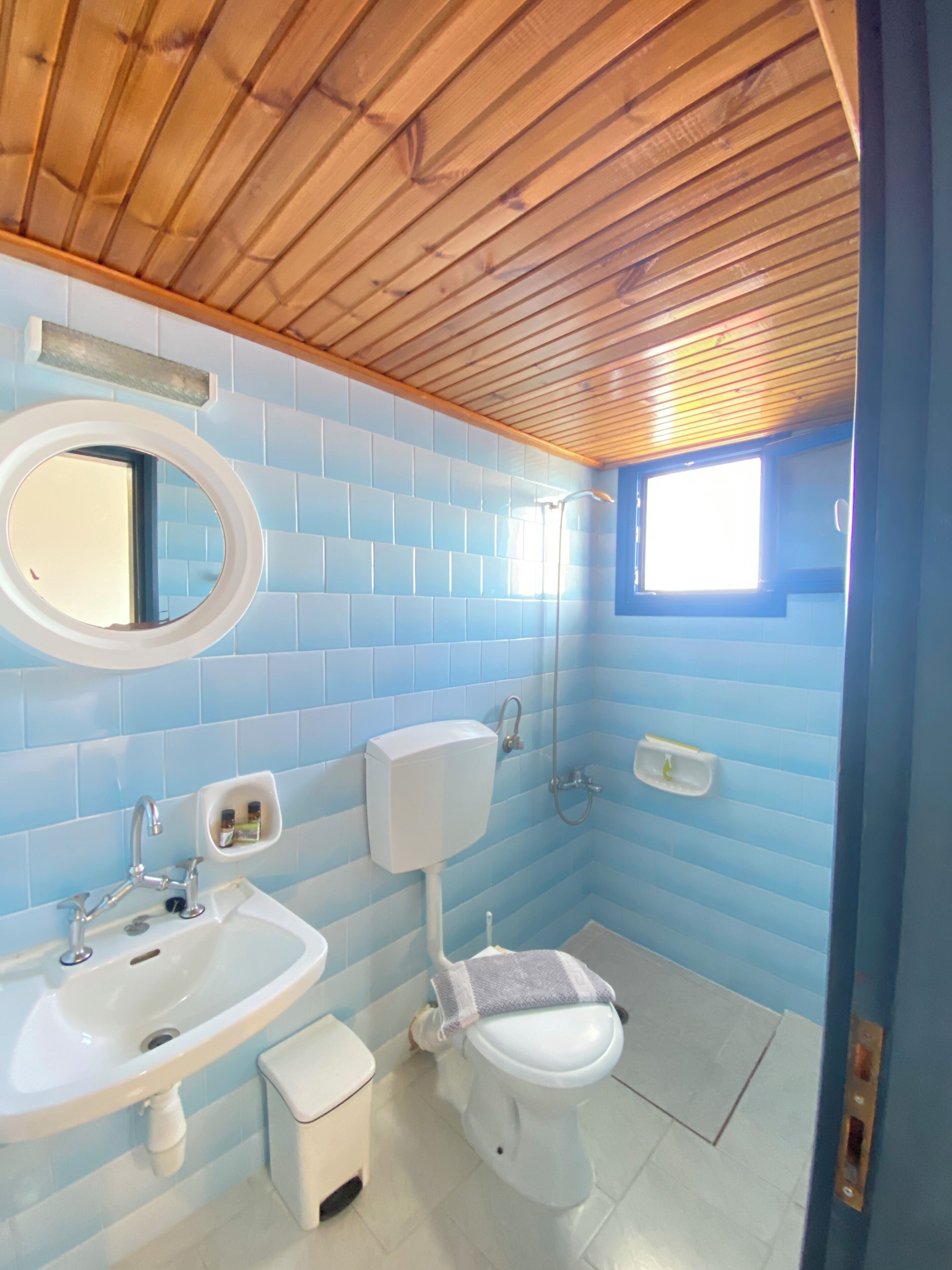 Un baño con azulejos azules, techo de madera, lavamanos, inodoro y ventana.