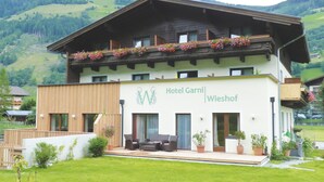 Een hotel met twee verdiepingen, modern van ontwerp, met een balkon met bloemen, een terras met buitenmeubilair en uitzicht op de omliggende heuvels.