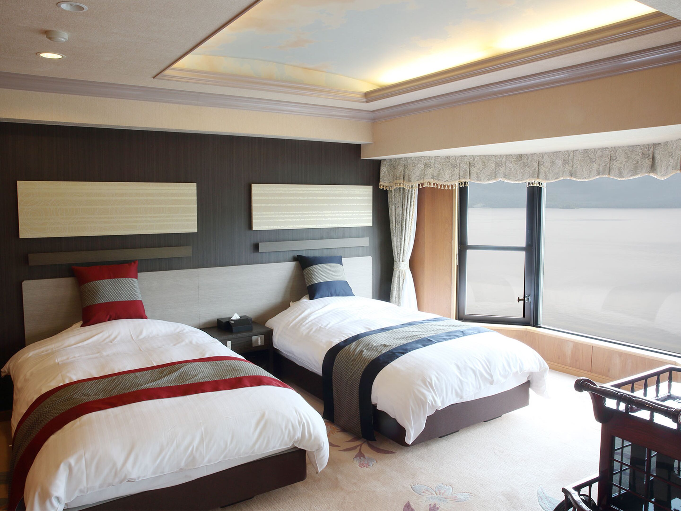 A hotel room with two beds, a nightstand, a window with curtains, and a view of the beach.