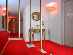 A hotel room with a red carpet, a gold-framed mirror, a console table with decorative items, and a door with a number sign. - Divani Ali Hotel (Istanbul)