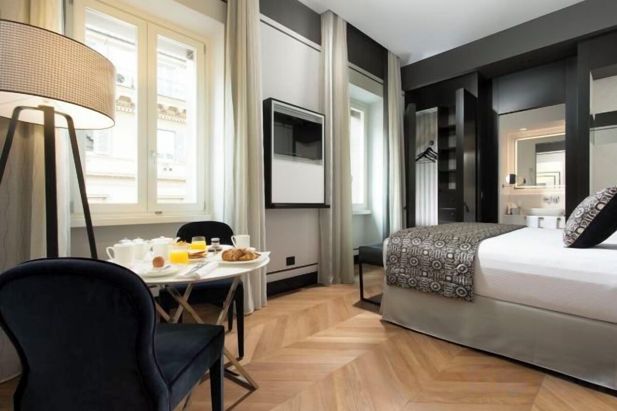 A hotel room with a bed, a TV mounted on the wall, a dining table with chairs, and a window with curtains.