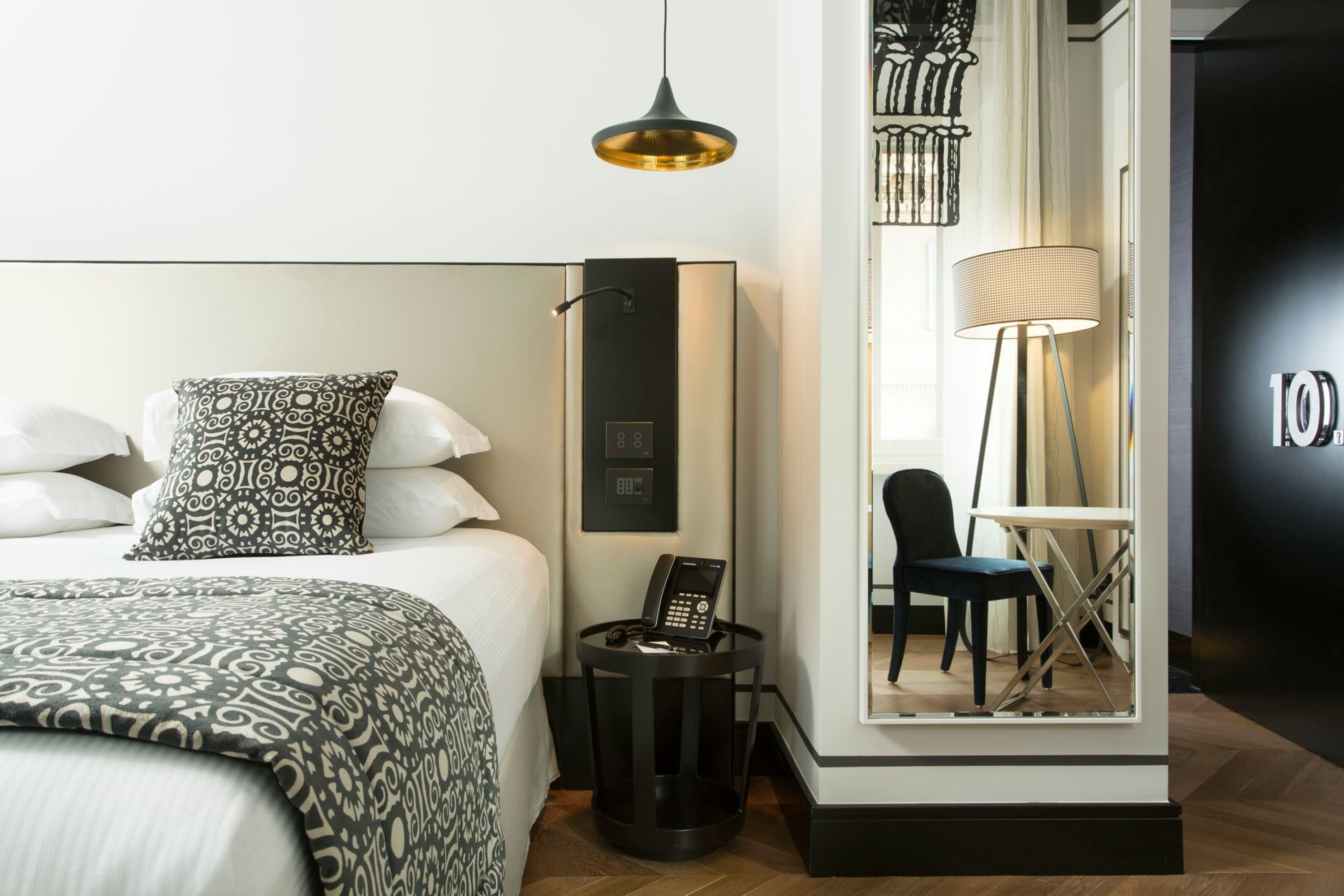 A modern hotel room with a large bed, patterned pillows, a bedside phone, a standing lamp, and a chair near a window.