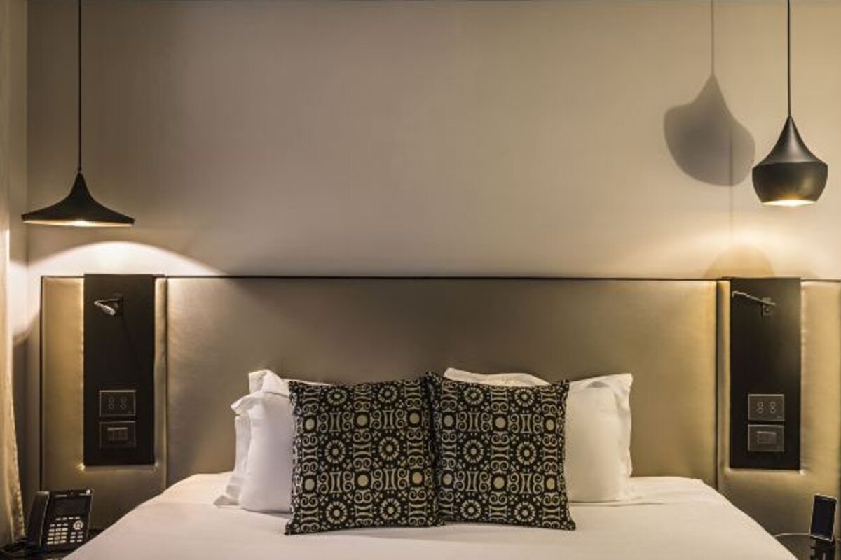 Une chambre d’hôtel avec un grand lit, deux coussins à motifs, un téléphone sur la table de chevet et deux lampes suspendues.