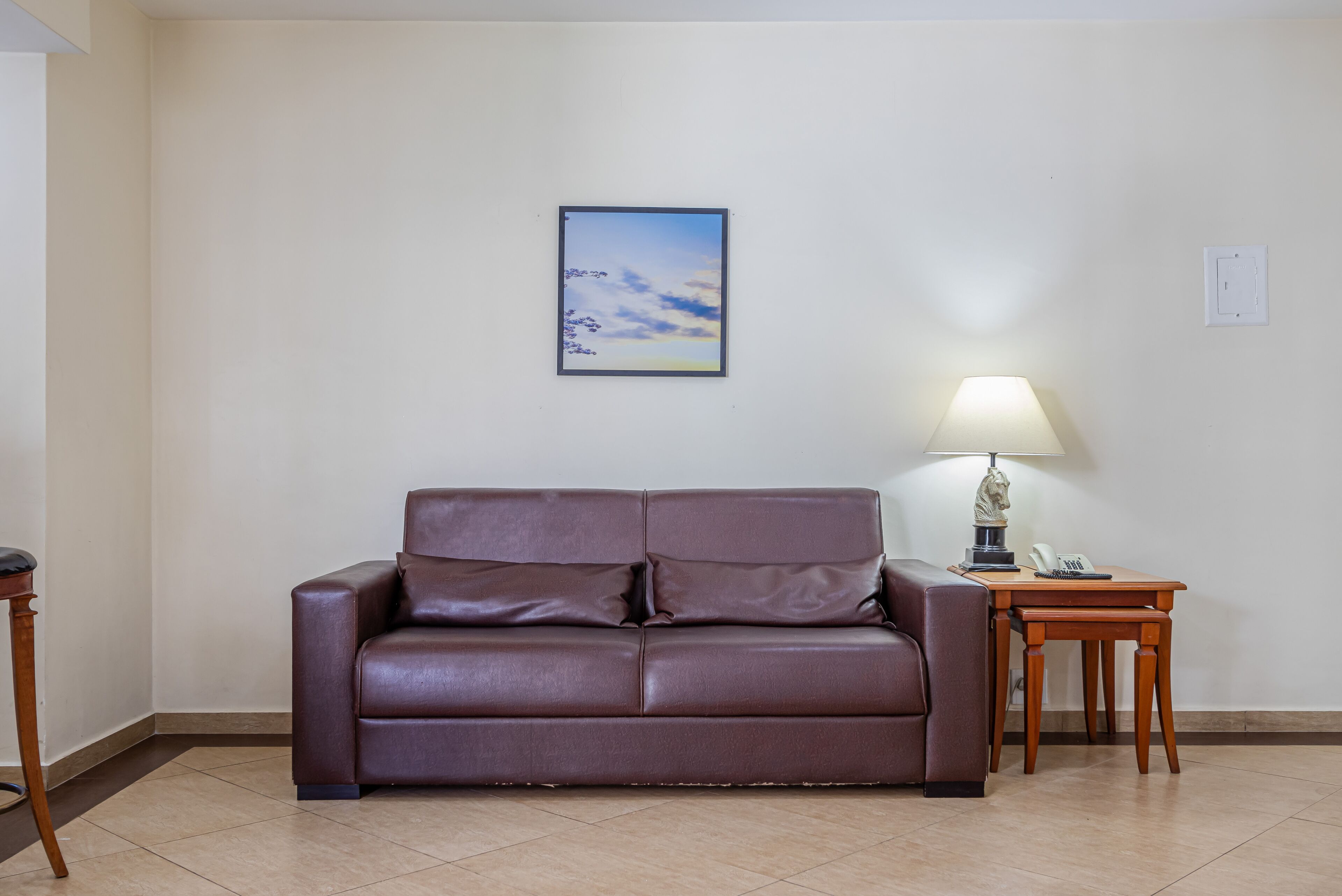 Um sofá de couro marrom encostado em uma parede branca, com um quadro emoldurado acima. À direita, uma mesa de madeira com um abajur e um telefone.