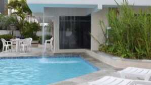 Una piscina con sillas reclinables blancas, una mesa y sillas, rodeada de vegetación y una fachada de edificio moderna.