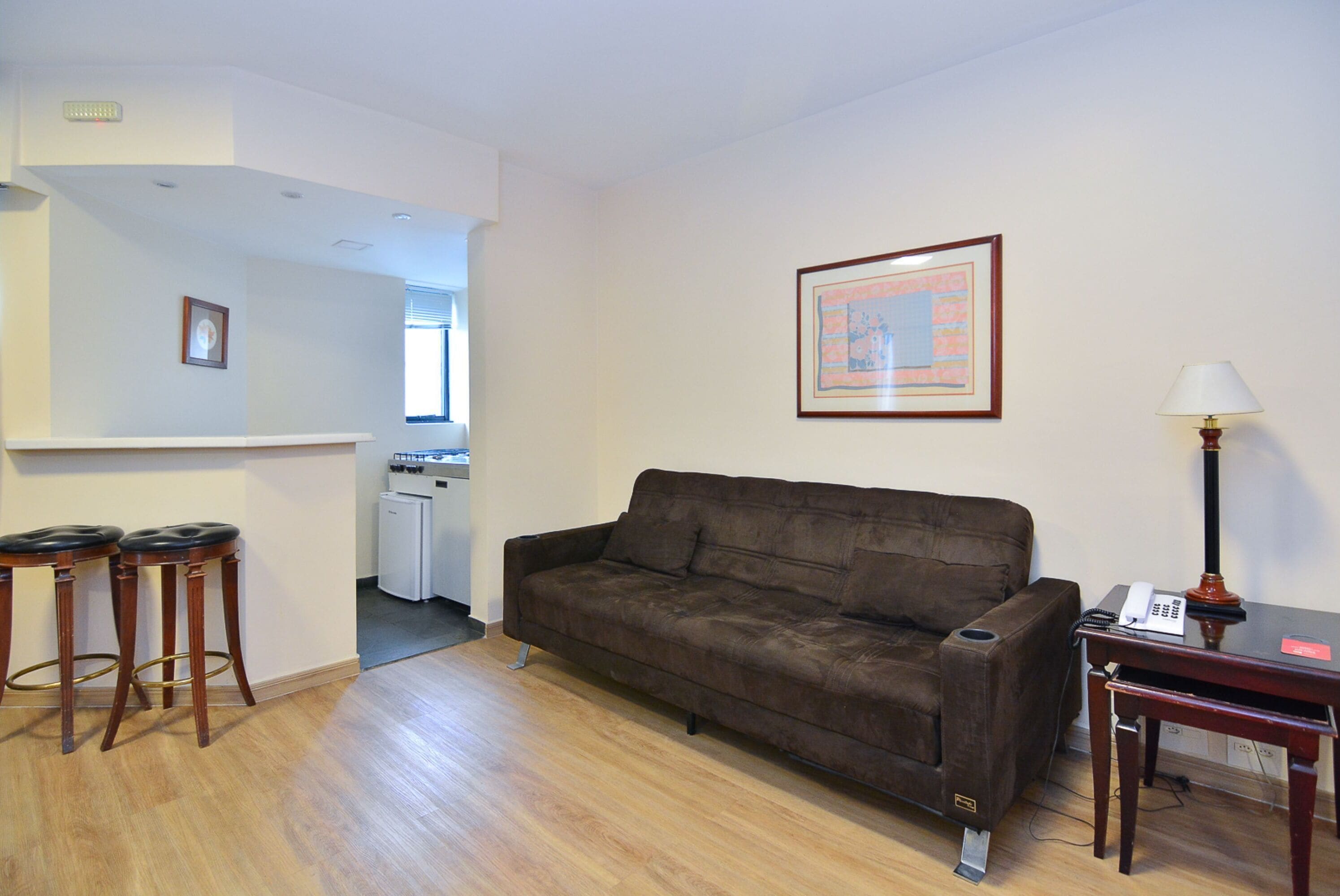 Ein Wohnzimmer mit braunem Sofa, Holzschemeln, einem kleinen Tisch mit Lampe und einem gerahmten Bild an der Wand.