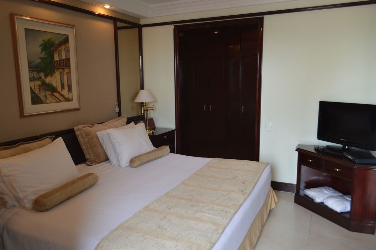 A hotel room with a bed, a television, a desk with a lamp, a wardrobe, and a painting on the wall.