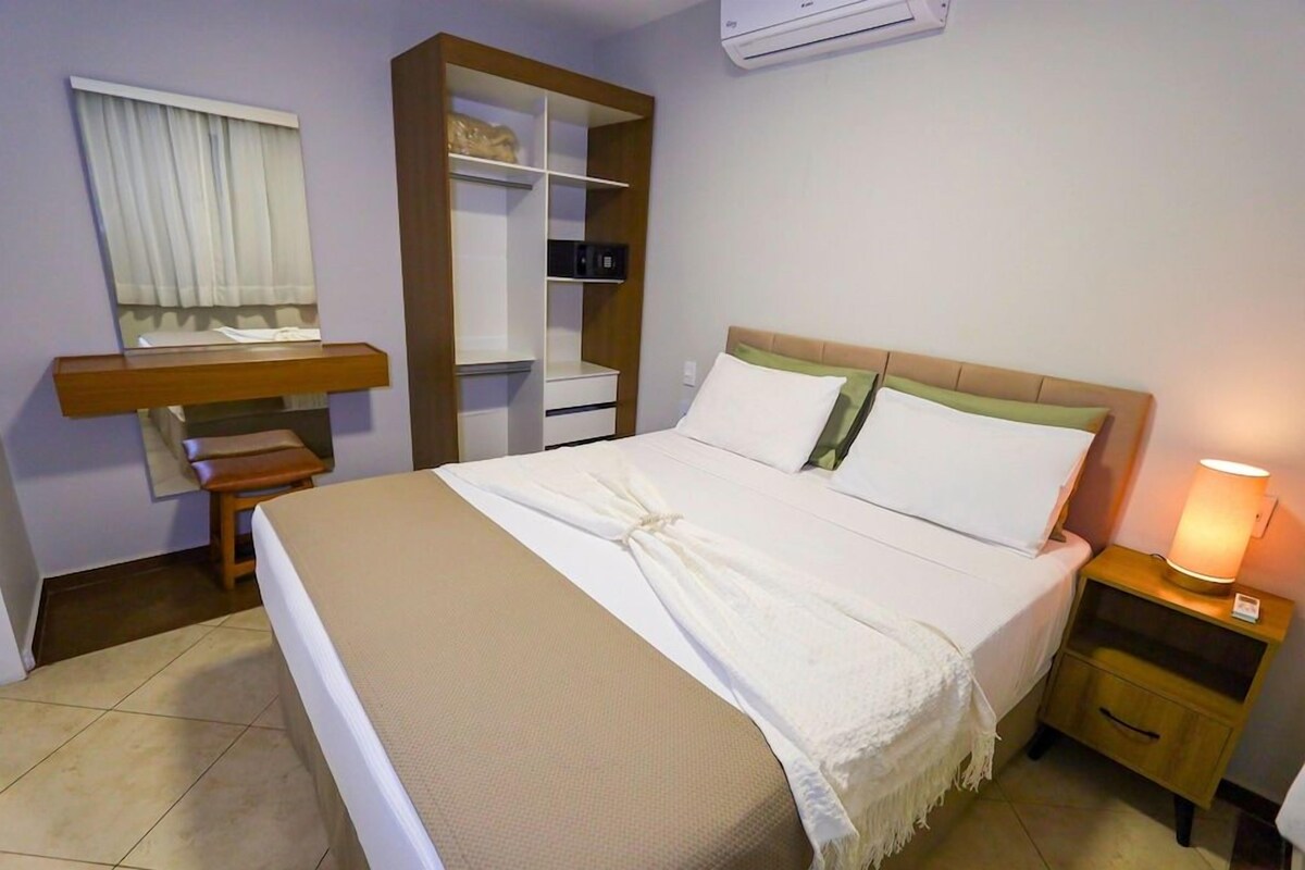 A hotel room with a bed, bedside table, nightstand, wardrobe, and a window with curtains.