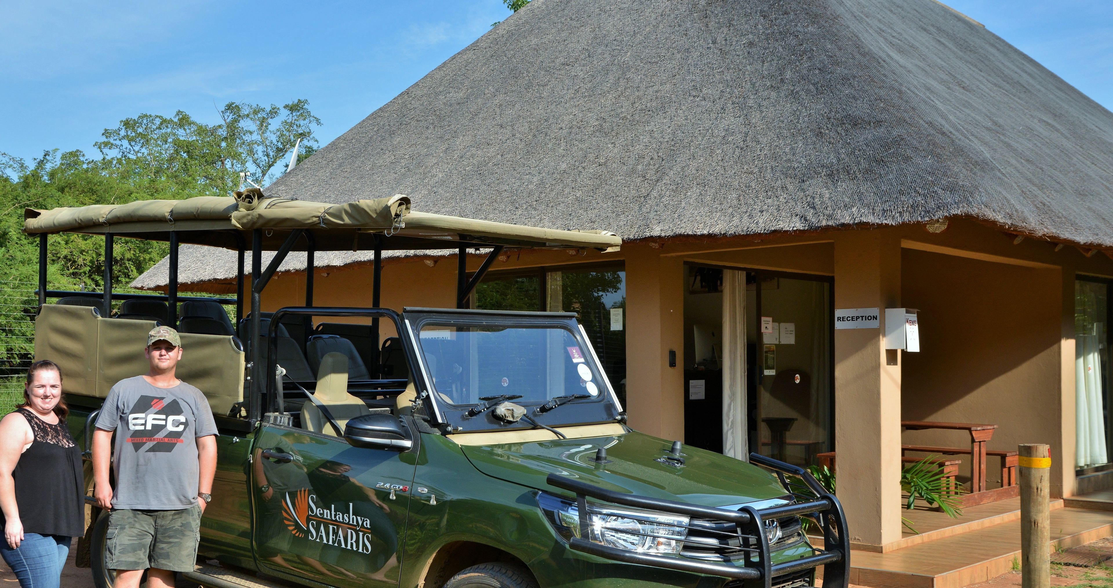 Een groene safari jeep met een luifel, geparkeerd voor een gebouw met een rieten dak.