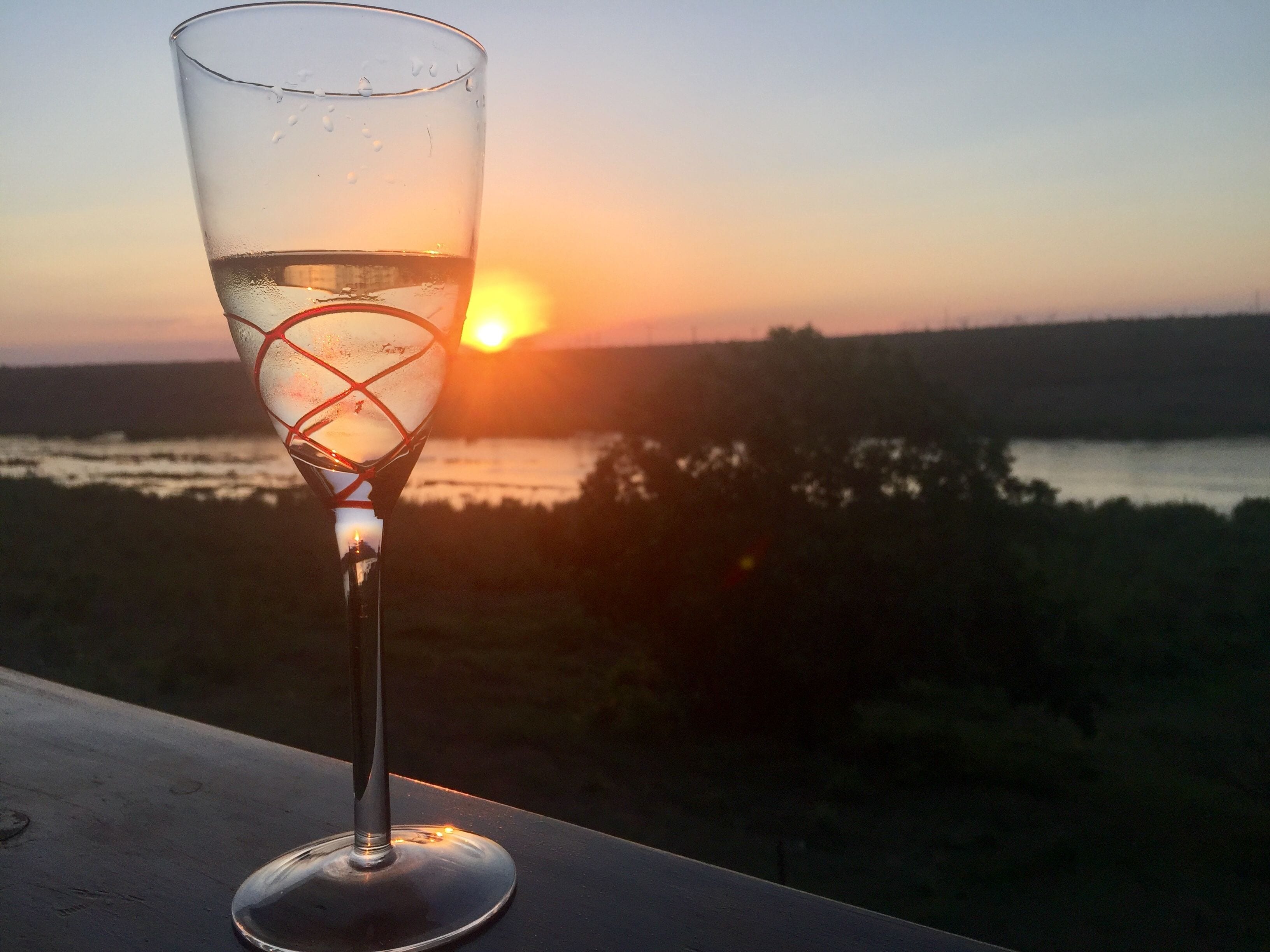 A glass of white wine on a balcony overlooking a sunset over a body of water.