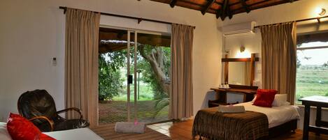 A hotel room with a large sliding glass door leading to a garden, a bed with a red pillow, a desk with a mirror, and a chair.