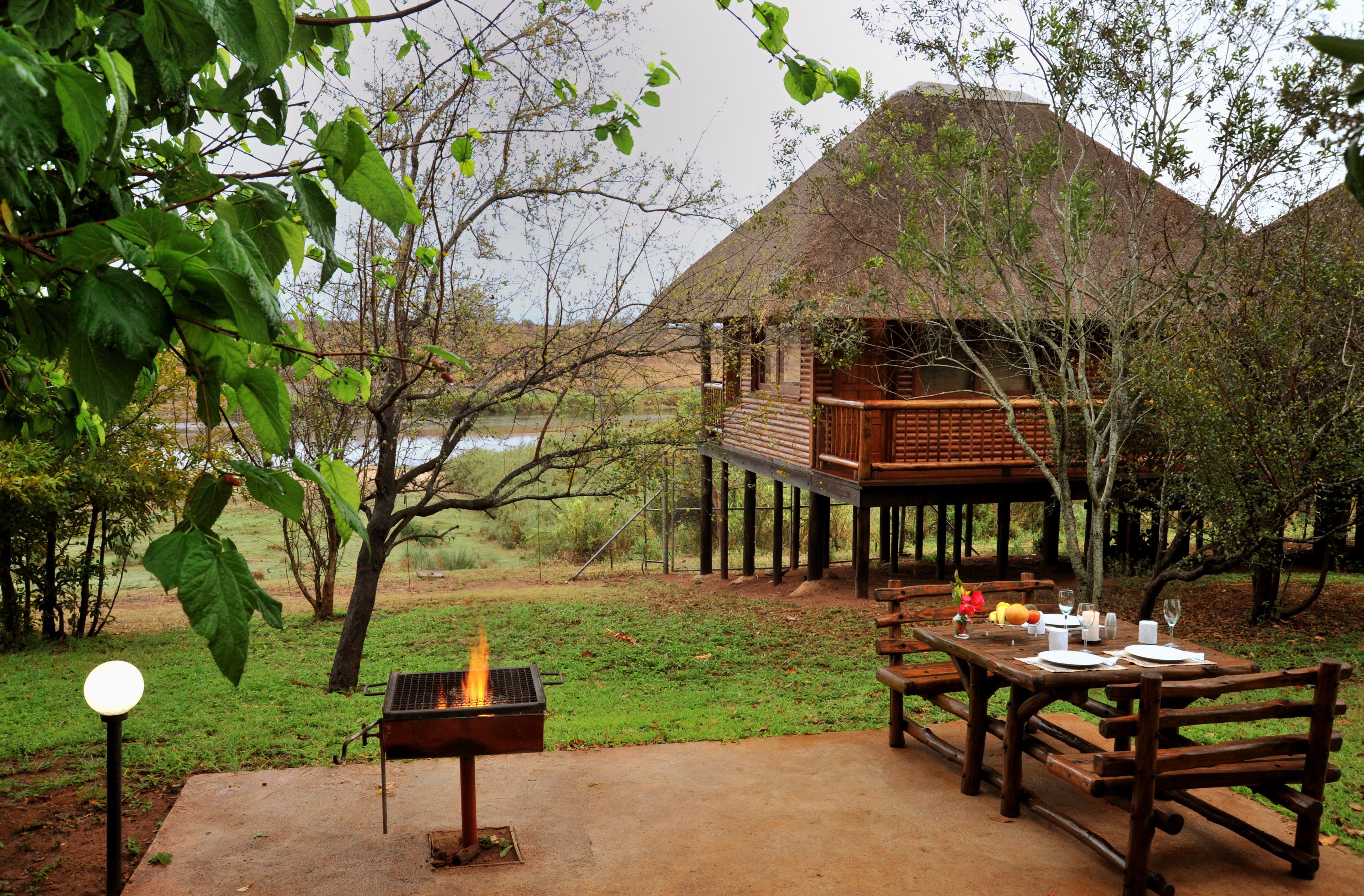 A wooden deck with a grill, dining table, and benches, overlooking a body of water and a thatched-roof building.
