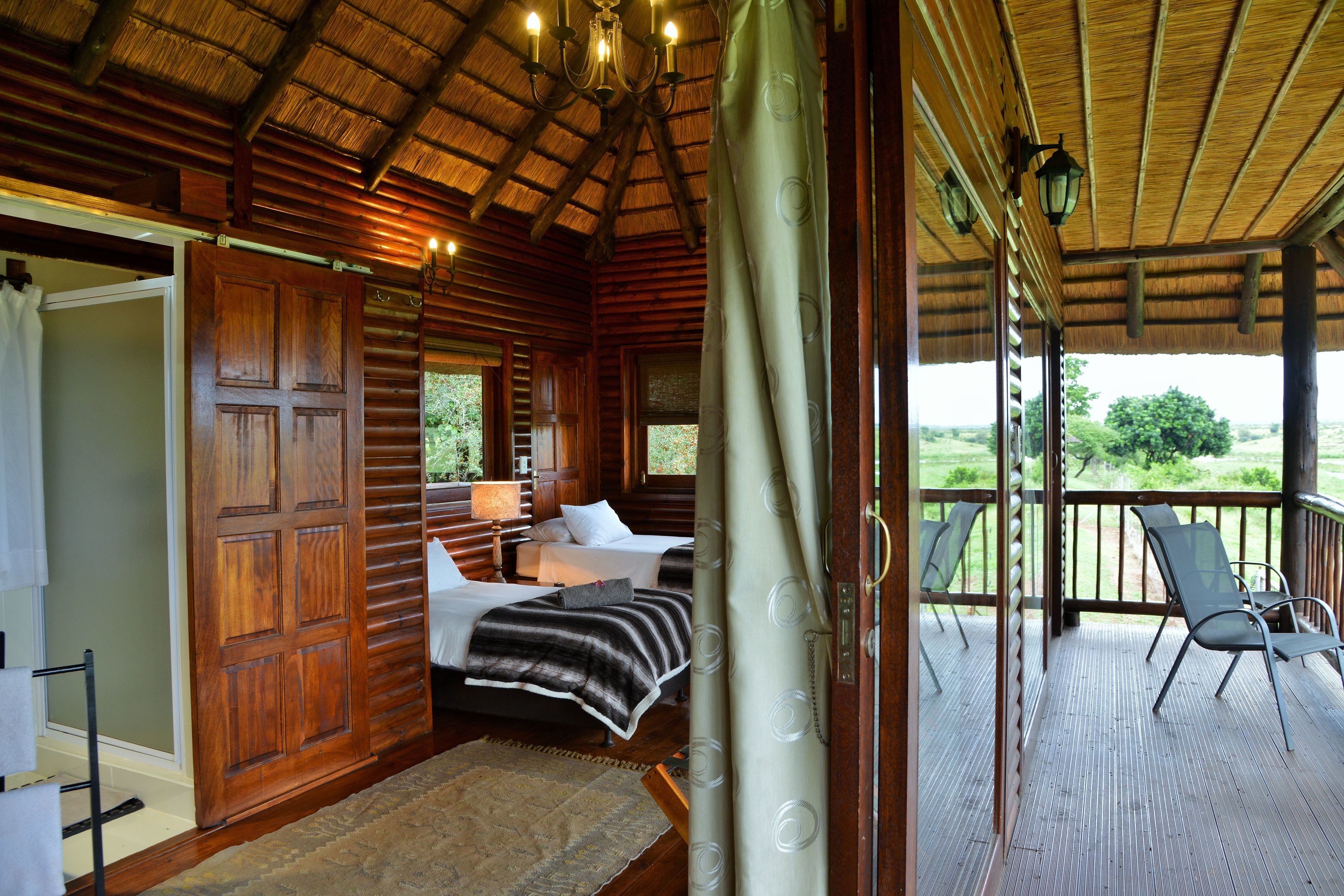 A wooden cabin with a thatched roof, a balcony with chairs, and a bed with a striped blanket.