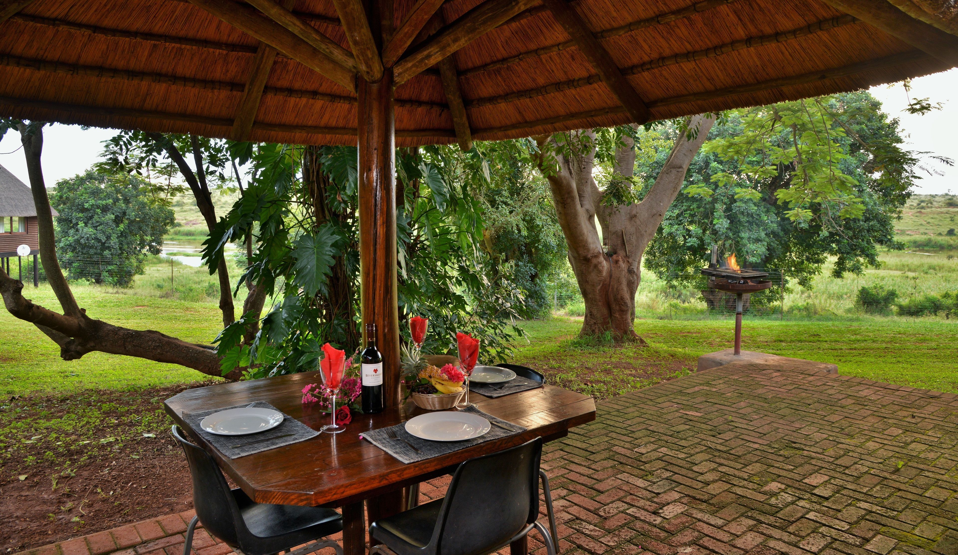 An outdoor dining area with a wooden table set for two, surrounded by lush greenery and trees.