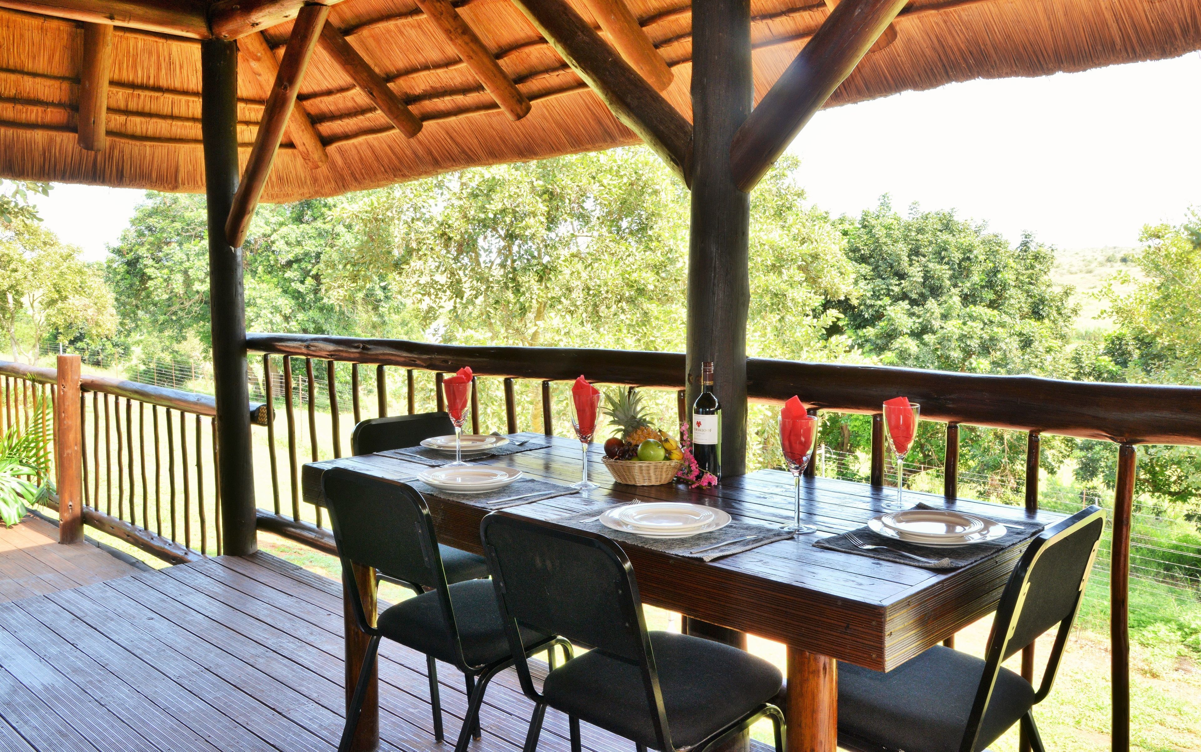 An outdoor dining area with a wooden table set for two, surrounded by chairs, under a thatched roof.