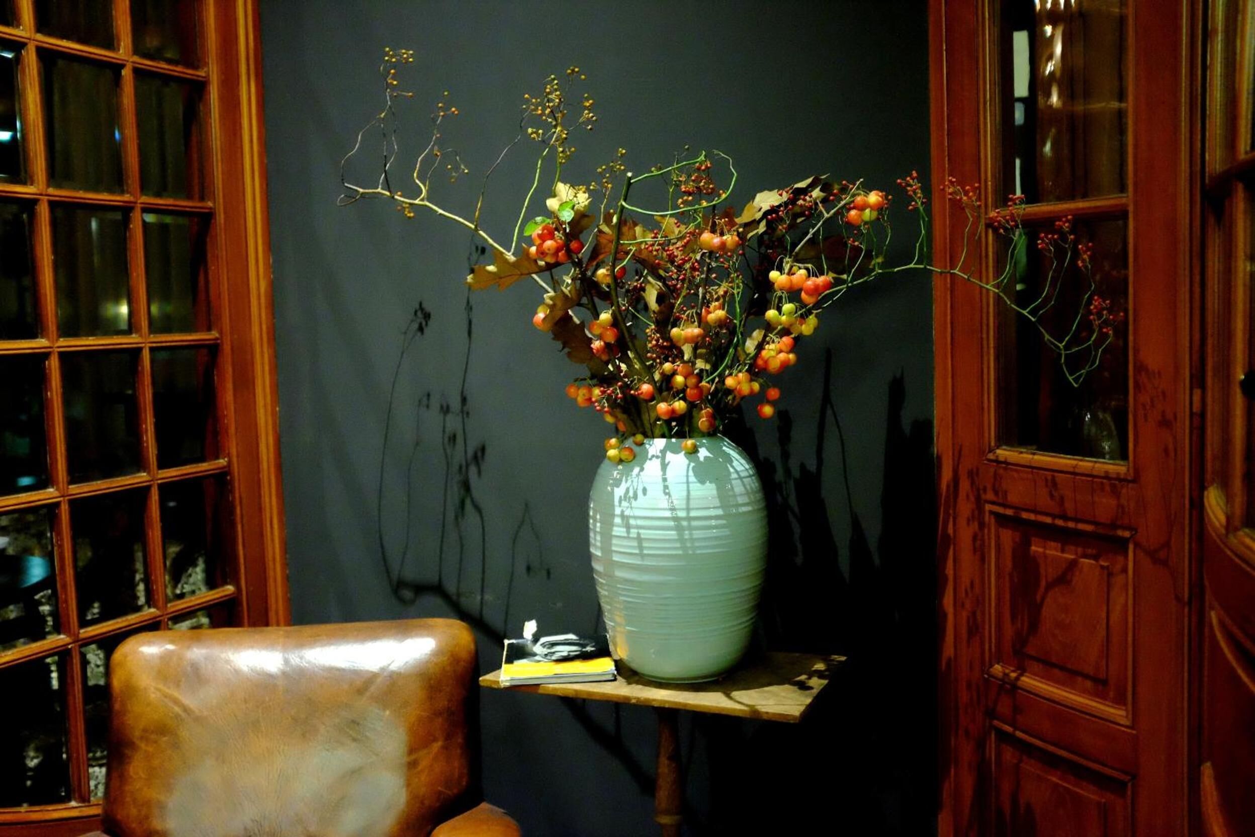 Une pièce avec une chaise en bois, une petite table sur laquelle est posé un vase de fleurs et un mur sombre.