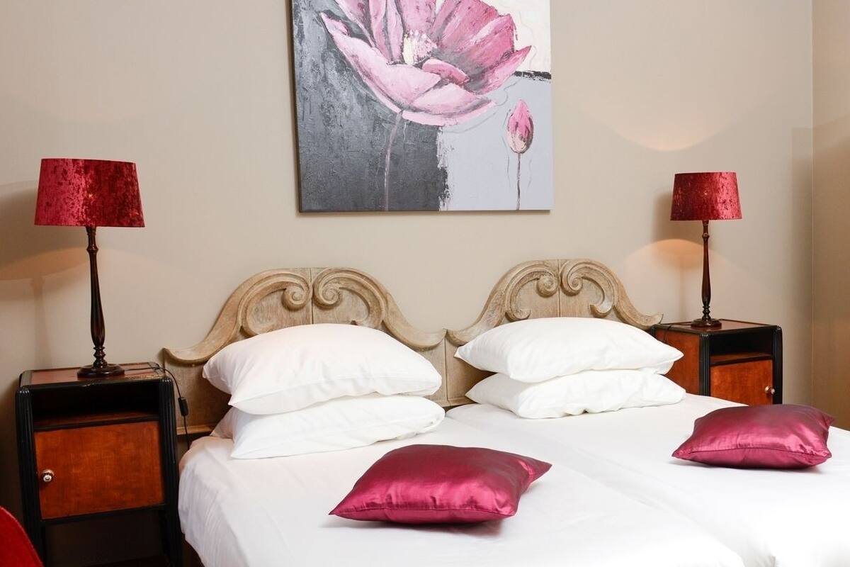 A double bed with white and pink pillows, two bedside tables with lamps, a wooden wardrobe, and a floral painting on the wall.