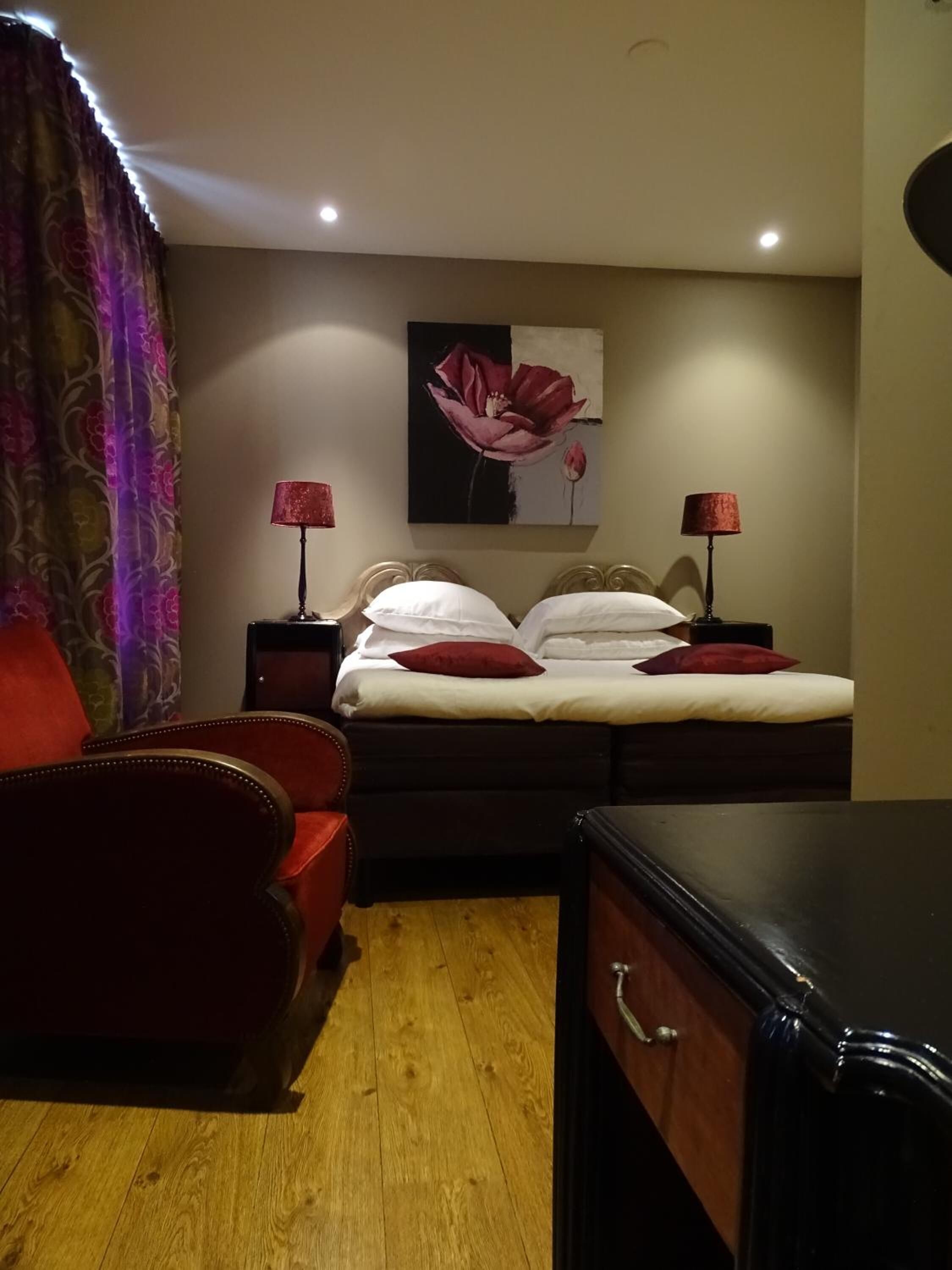 Ein Schlafzimmer mit einem Bett, zwei Nachttischlampen, einem roten Sessel, Holzboden und einem Gemälde an der Wand.