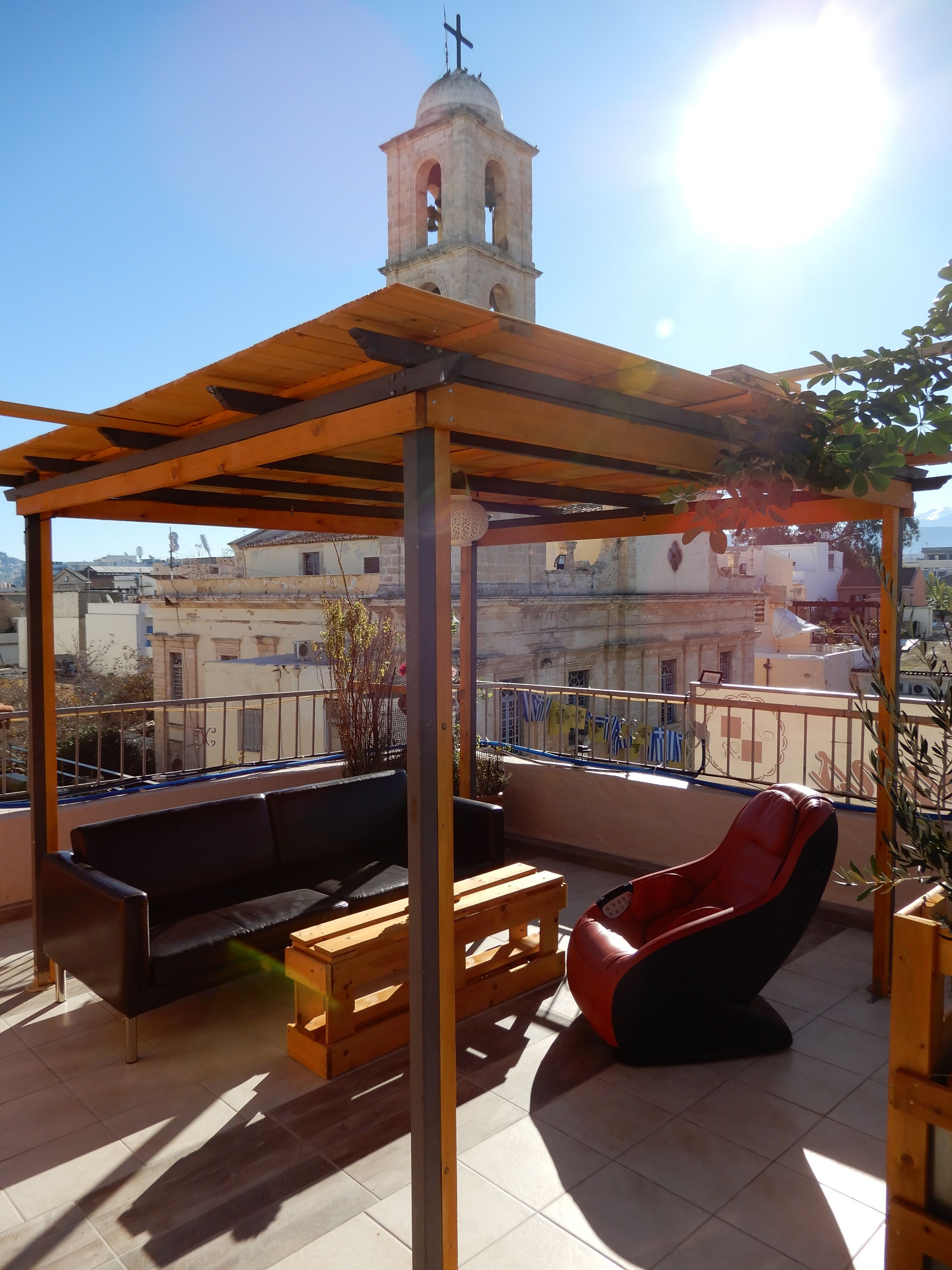 A rooftop patio with a wooden pergola, a black sofa, a red chair, and a wooden table. A bell tower is visible in the background.