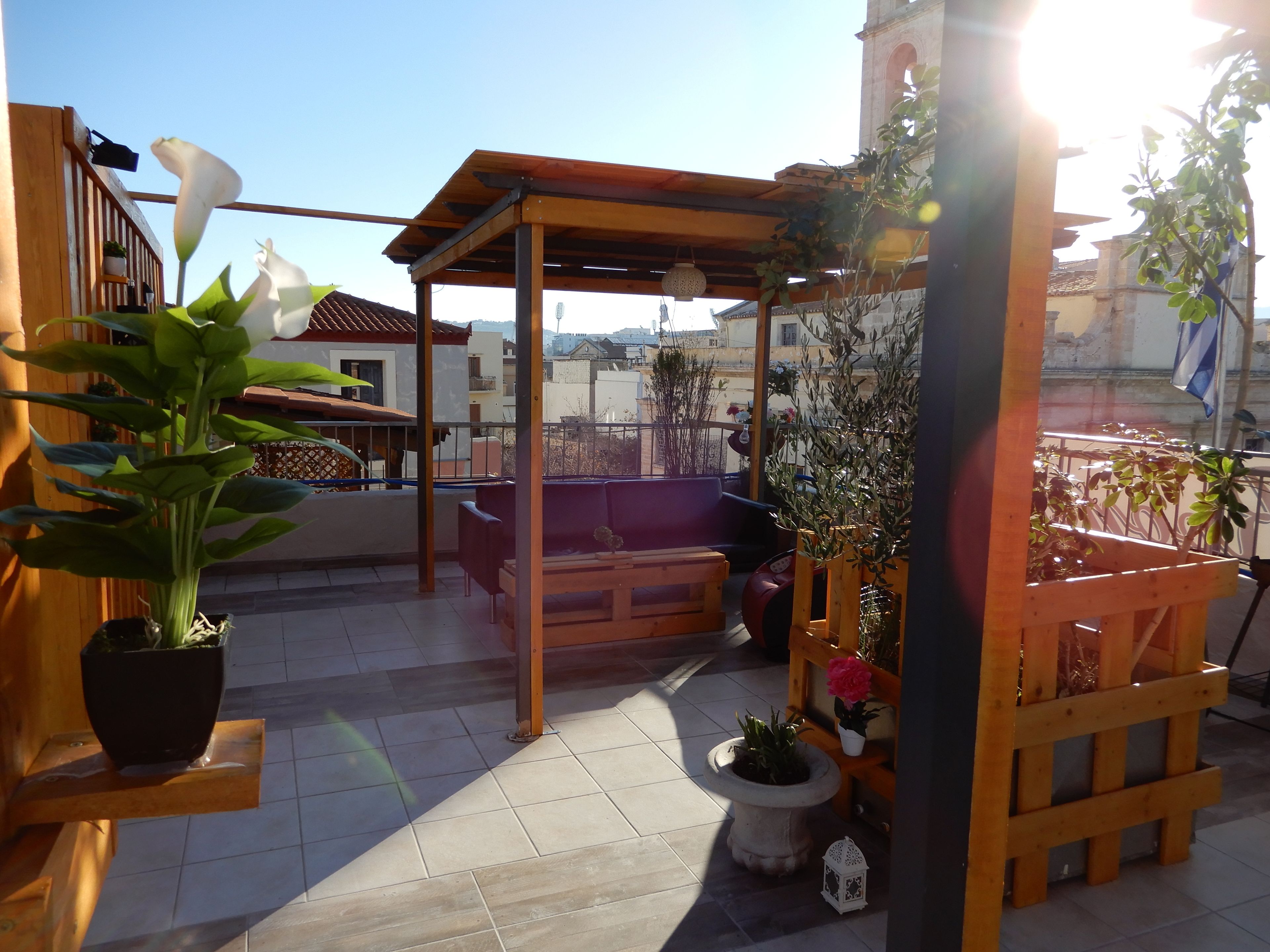 A rooftop terrace with a wooden pergola, seating area, potted plants, and a view of the cityscape.