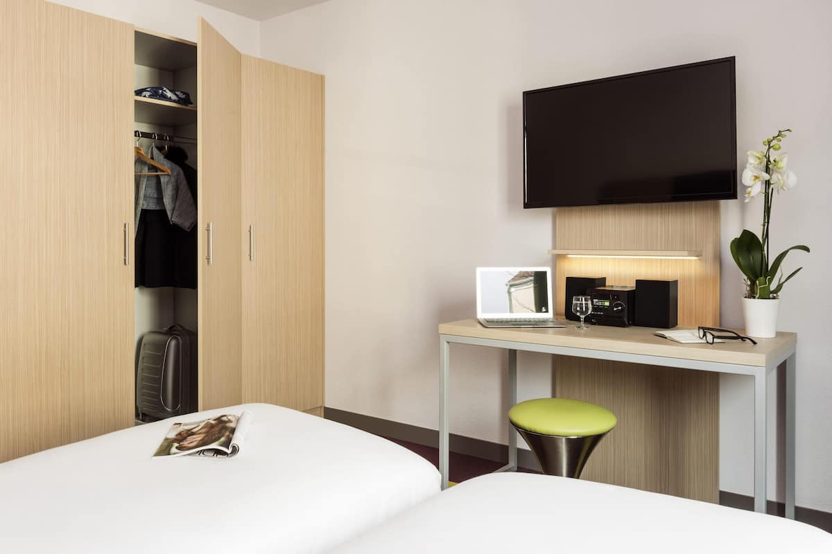 A hotel room with a bed, a desk with a laptop, a flat-screen TV, a potted orchid, and a wardrobe.