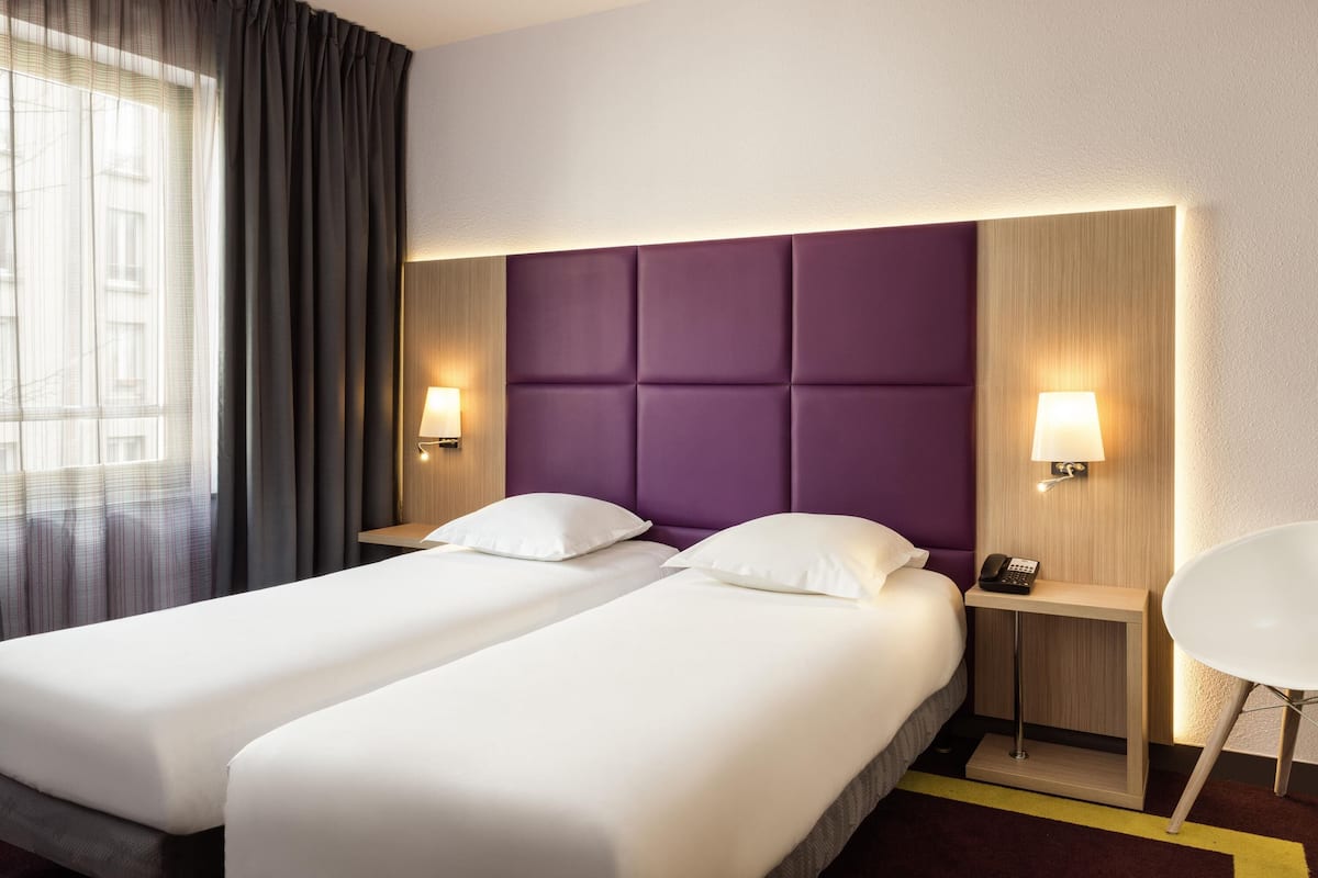 Ein Hotelzimmer mit zwei Betten, einem Nachttisch mit Telefon, einem Sessel und einem Fenster mit Vorhängen.
