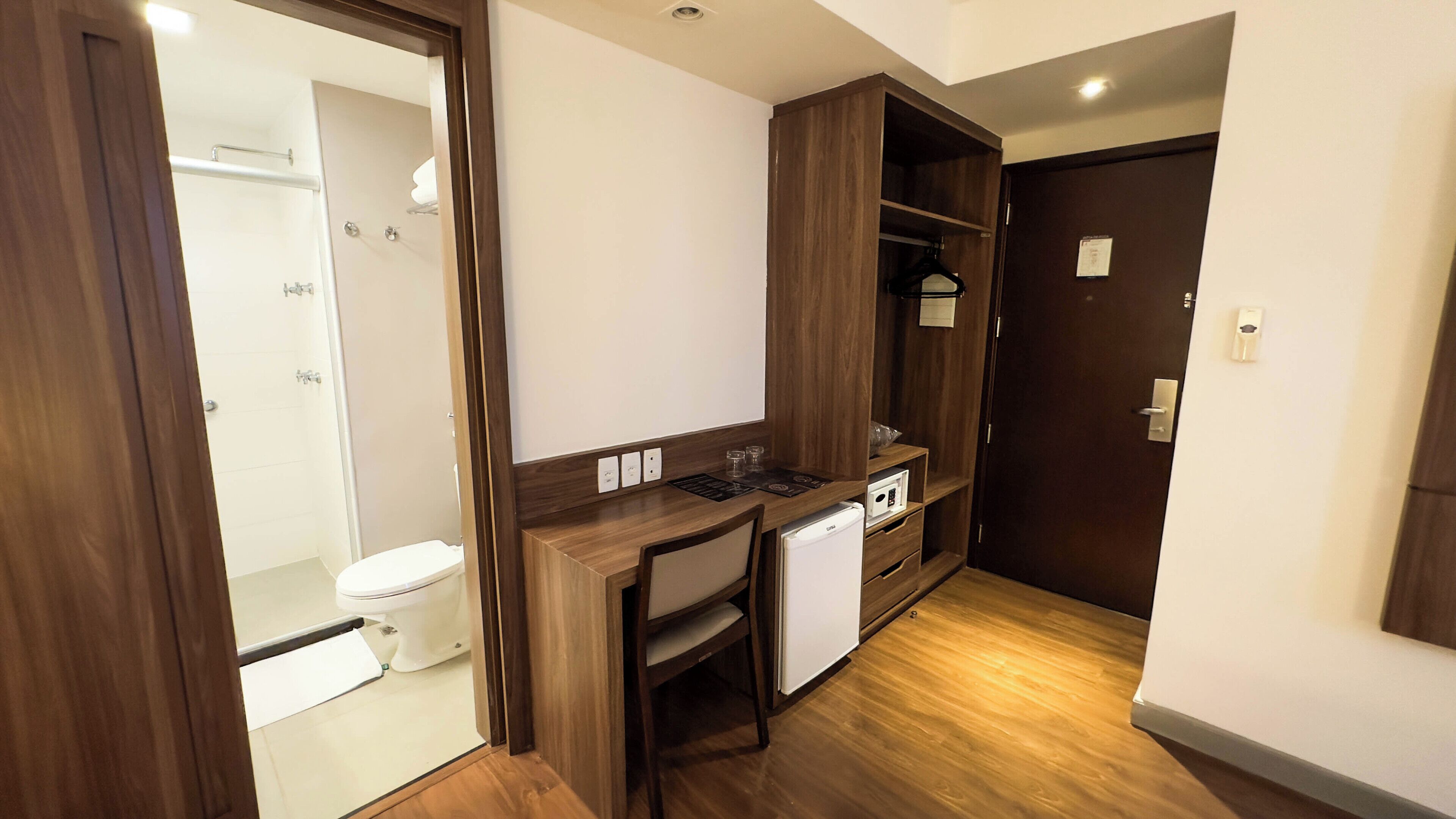 A compact bathroom with a toilet and shower, a wooden desk with a chair, and a small refrigerator.