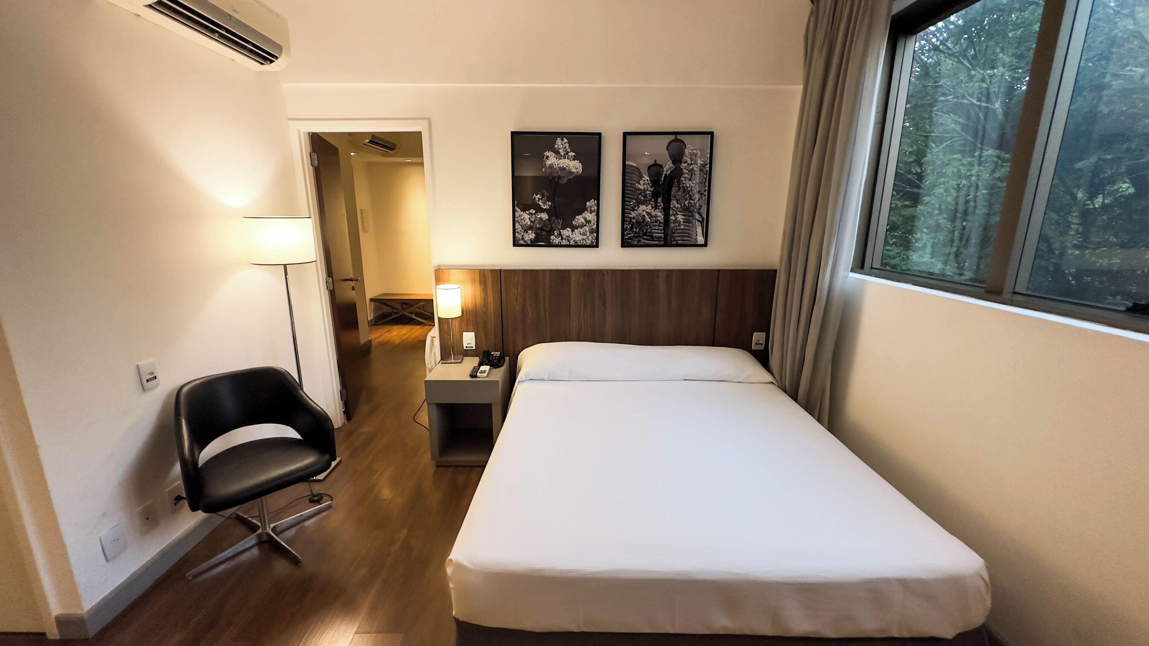 Habitación de hotel con una cama individual, un escritorio con lámpara, una silla y dos cuadros en la pared.