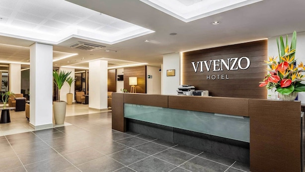 Una moderna zona de recepción de hotel con un mostrador de madera, un separador de vidrio y un gran arreglo floral.