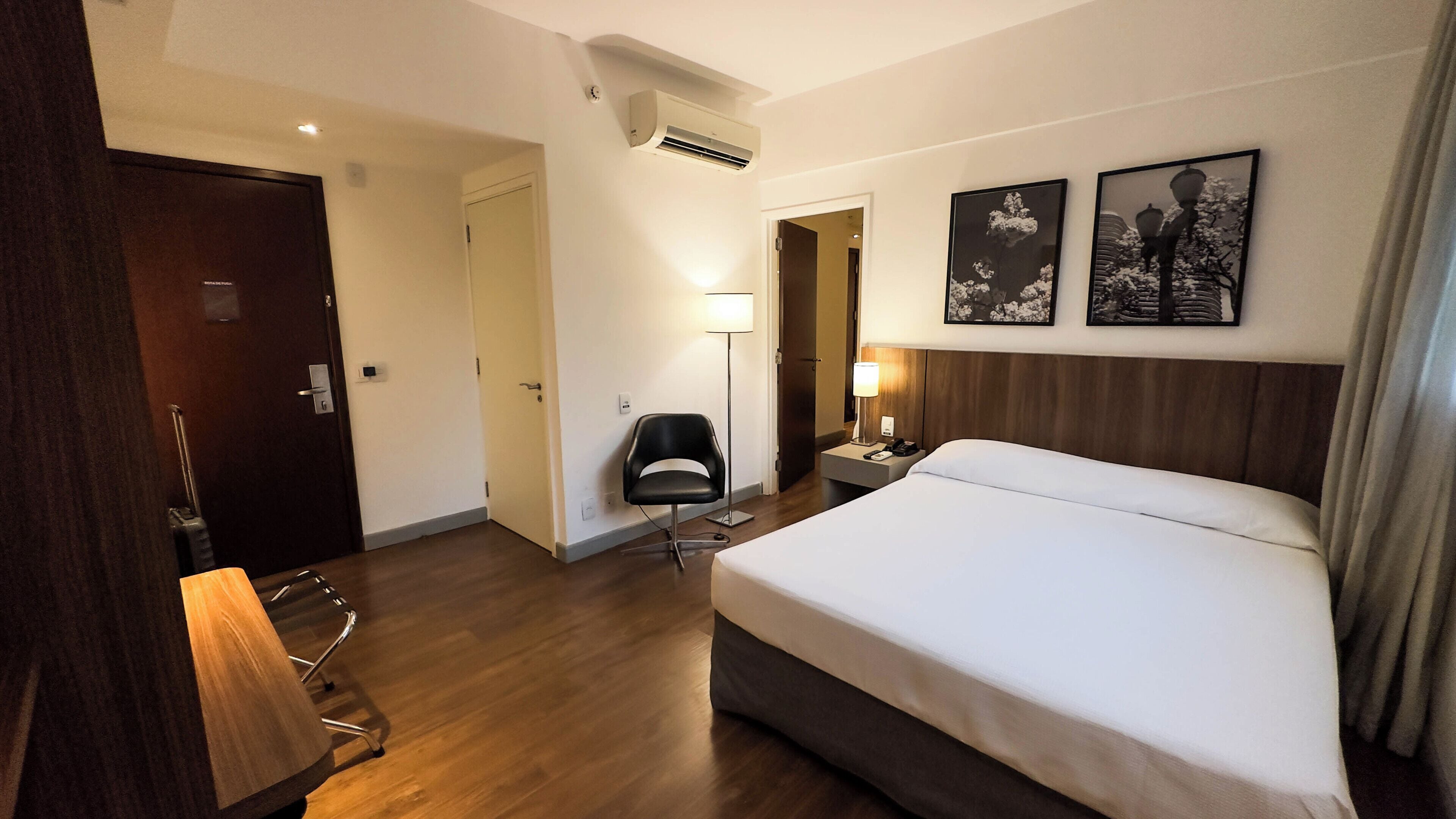 Une chambre d’hôtel avec un grand lit, un bureau avec une chaise, un lampadaire et deux tableaux encadrés au mur.