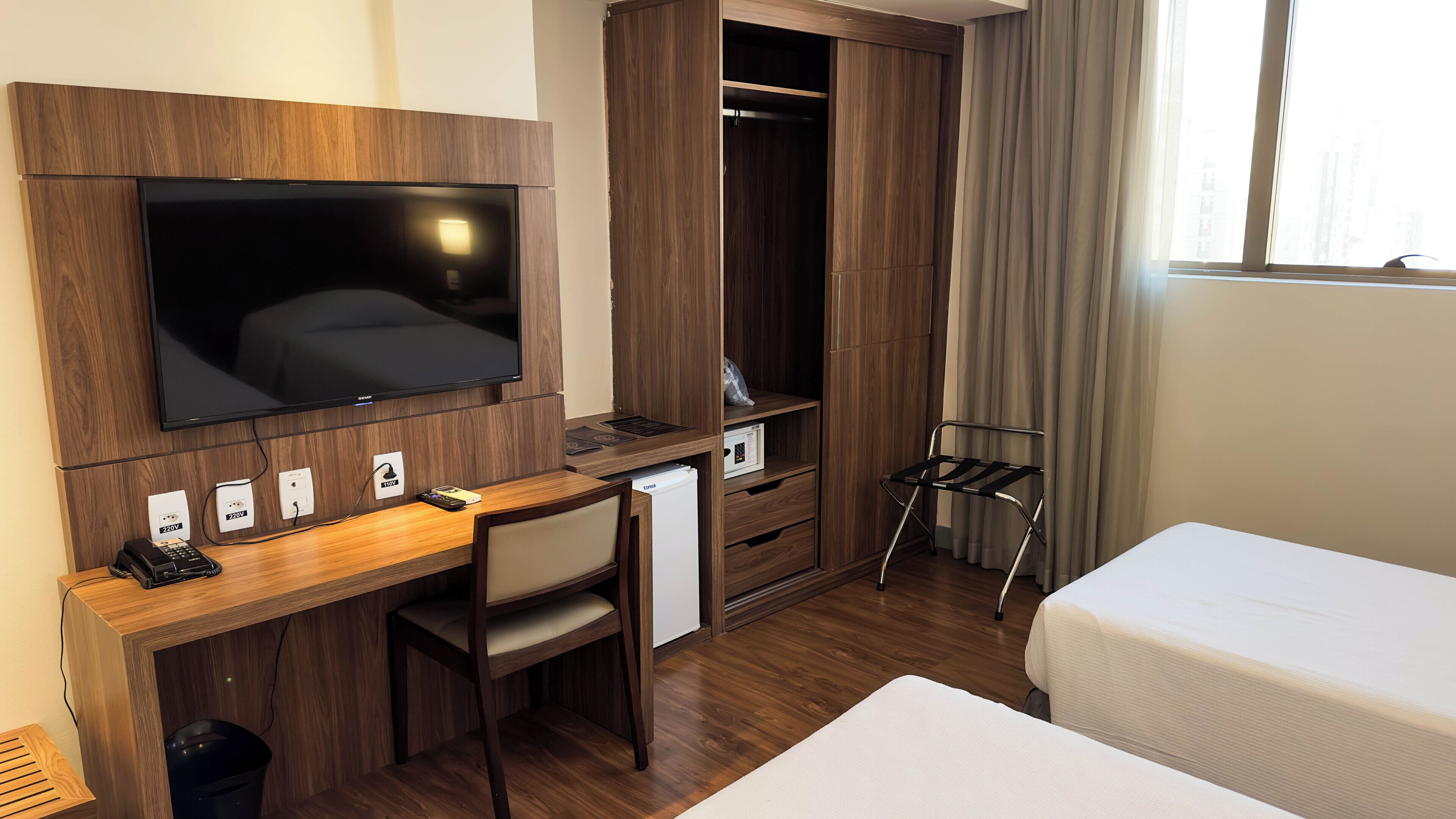 A hotel room with a wooden desk, a TV, a wardrobe, a bed, and a window with curtains.
