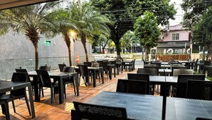 Un área de comedor al aire libre con mesas y sillas, rodeada de árboles y un muro de piedra.