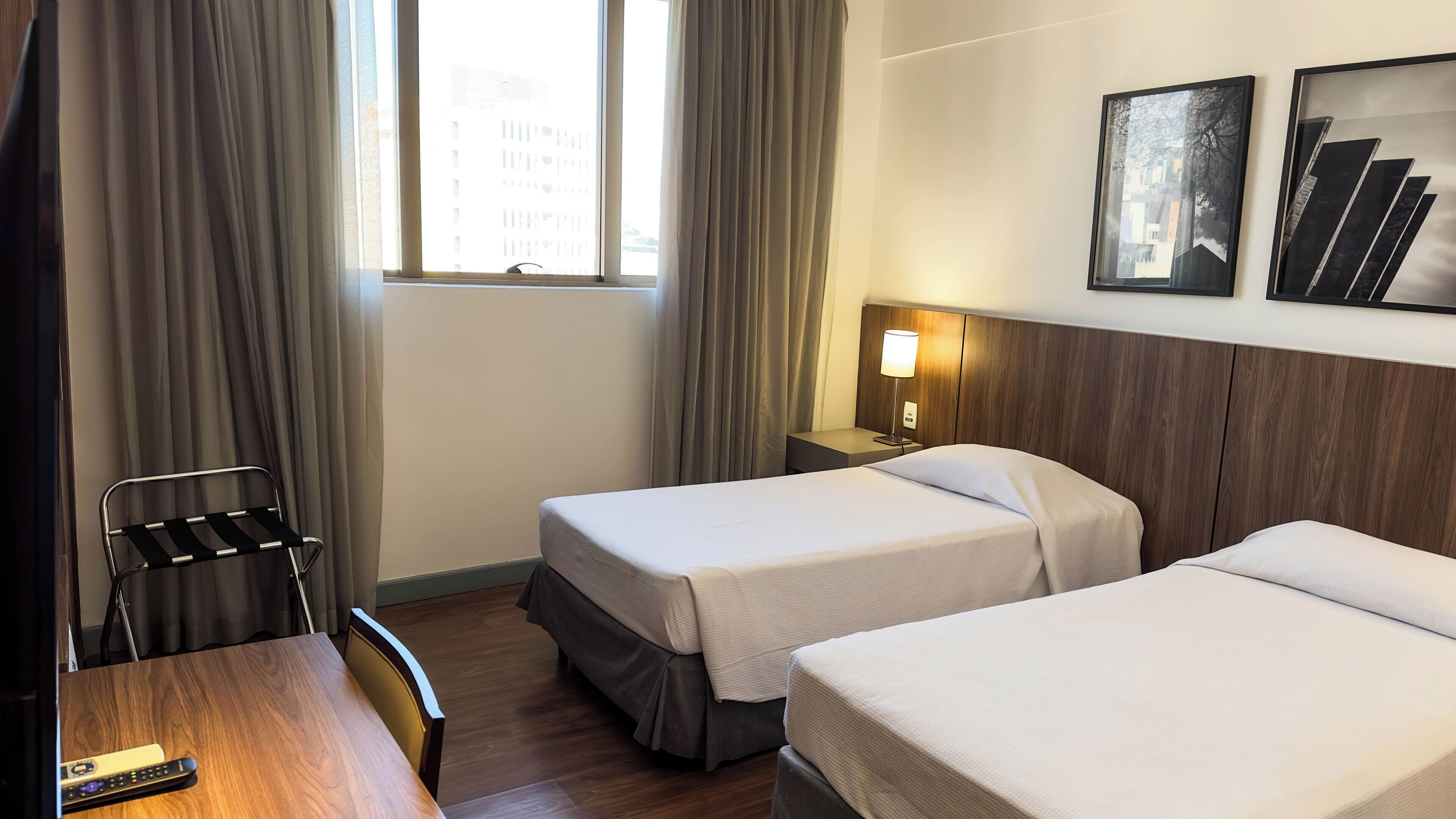 A hotel room with two single beds, a wooden desk, a chair, a nightstand with a lamp, and a window with curtains.