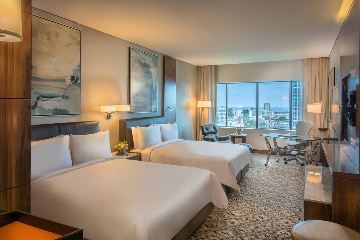 Habitación de hotel con dos camas, un escritorio, una silla y un amplio ventanal con vistas a la ciudad.