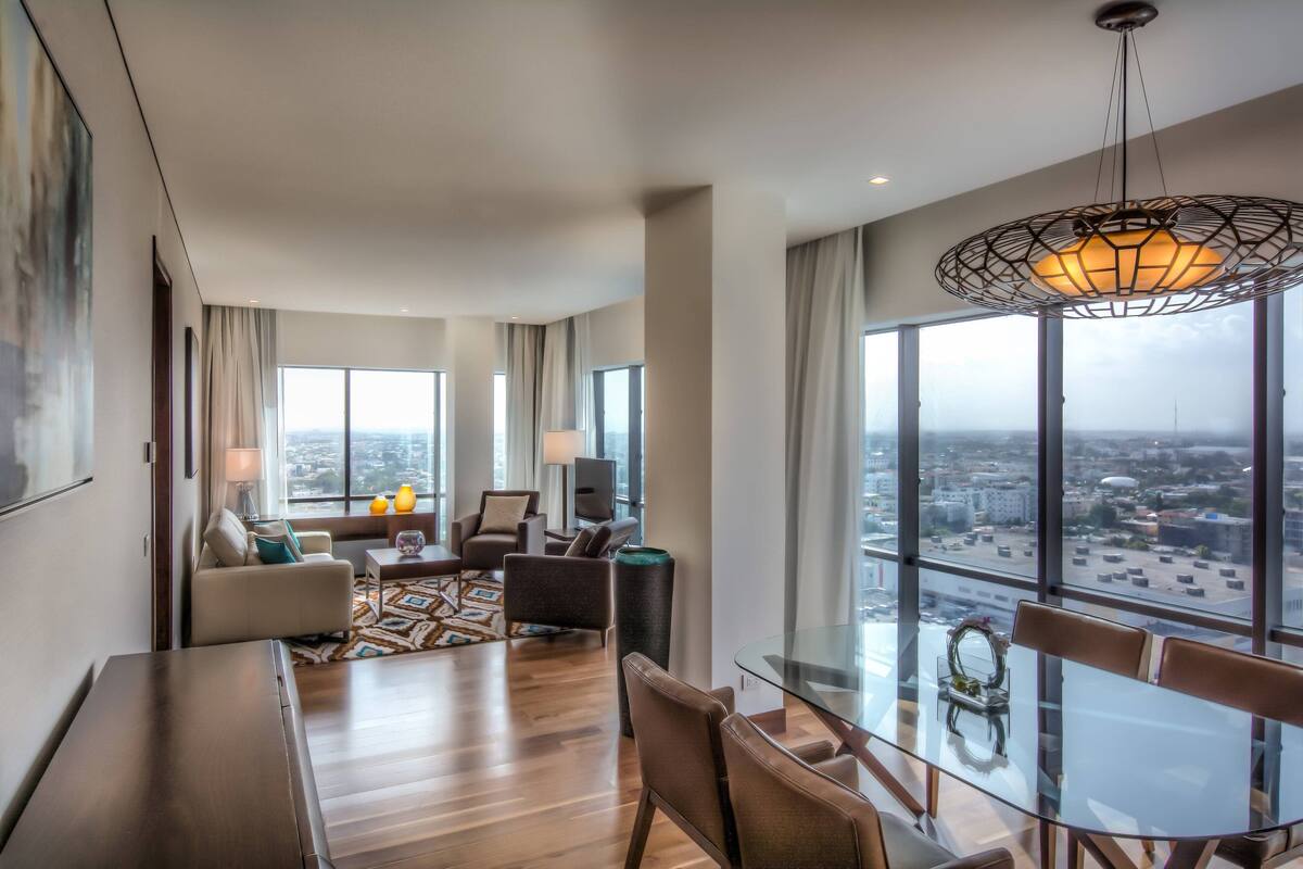 Una habitación de hotel moderna con zona de comedor, ventanas amplias y vista a la ciudad.