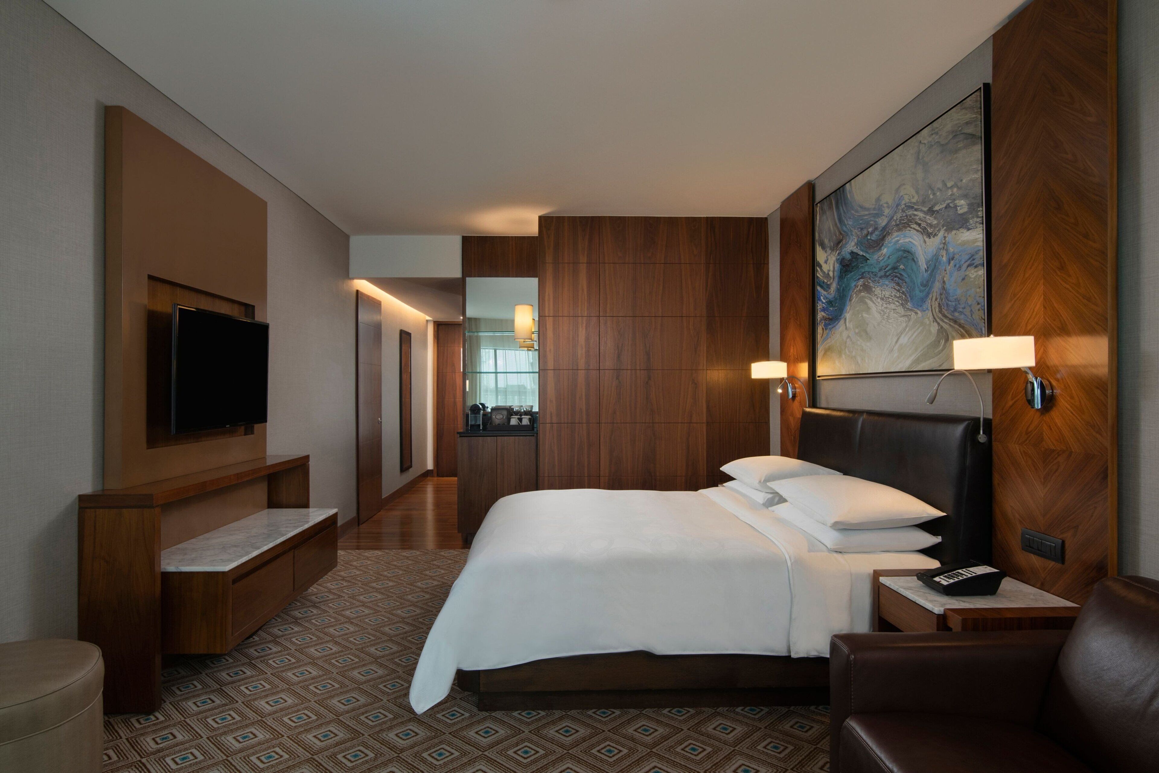Quarto de hotel com uma cama grande, uma televisão presa na parede, uma mesa com abajur e um quadro sobre a cama.