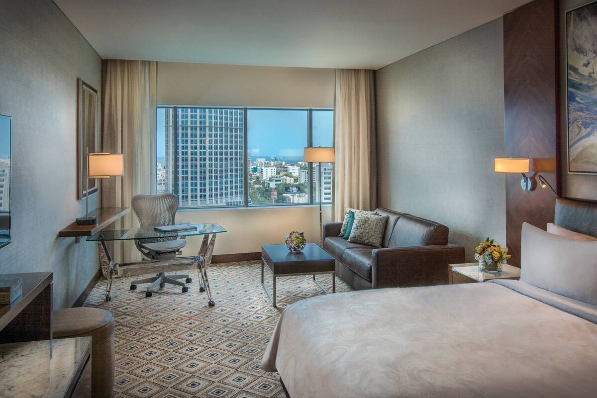 Habitación de hotel con cama, escritorio, silla, sofá y ventana con vista a la ciudad.