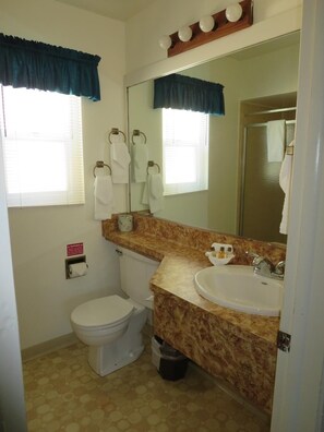 A bathroom with a toilet, sink, and mirror. There are towels hanging on hooks, a trash bin, and a warning sign on the wall. - Bridge Bay Lodge at Shasta Lake (Redding)