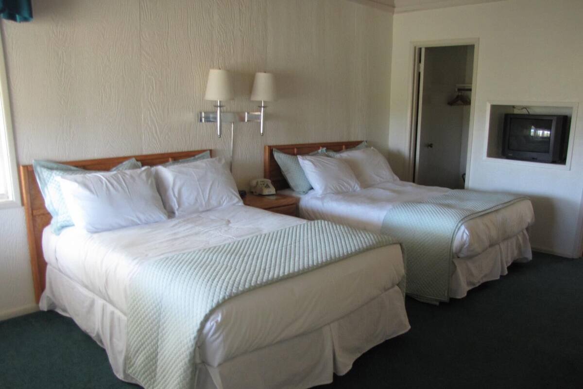 A hotel room with two beds, each with white linens and a wooden headboard. There are two wall-mounted lamps on the wall above the beds.