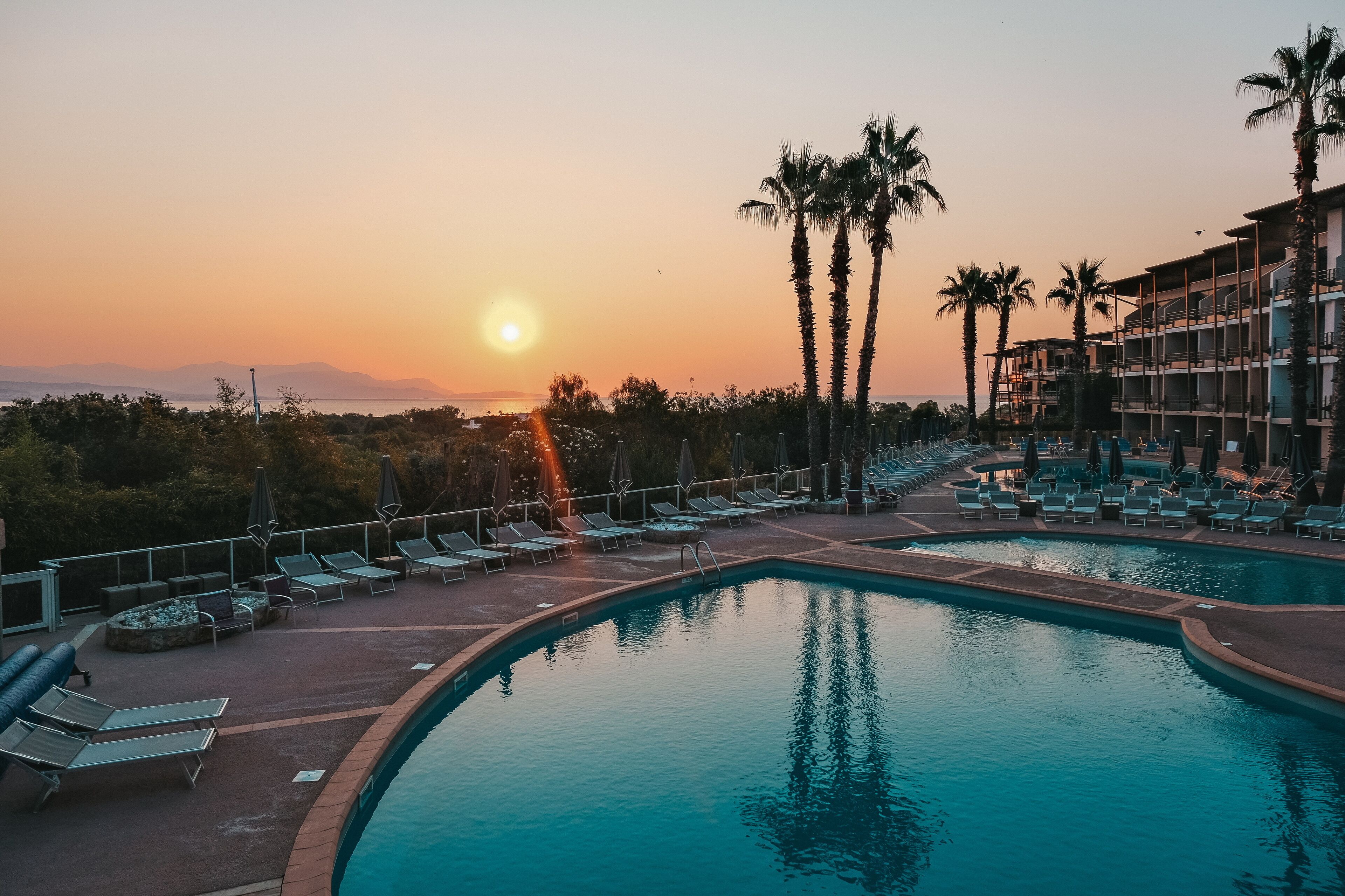 Ein Poolbereich eines Resorts mit Liegestühlen und Palmen, umgeben von einem Gebäude mit mehreren Balkonen.