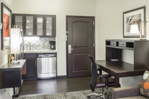 Une cuisine compacte dotée d’appareils électroménagers modernes, un coin repas avec une table en bois foncé et une chaise de bureau, et une porte ornée d’un panneau décoratif.