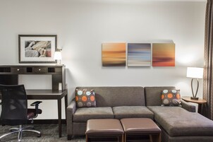 Una habitación de hotel moderna con un sofá gris, un escritorio y una silla. Hay tres obras de arte colgadas en la pared y una lámpara sobre una mesita de noche.